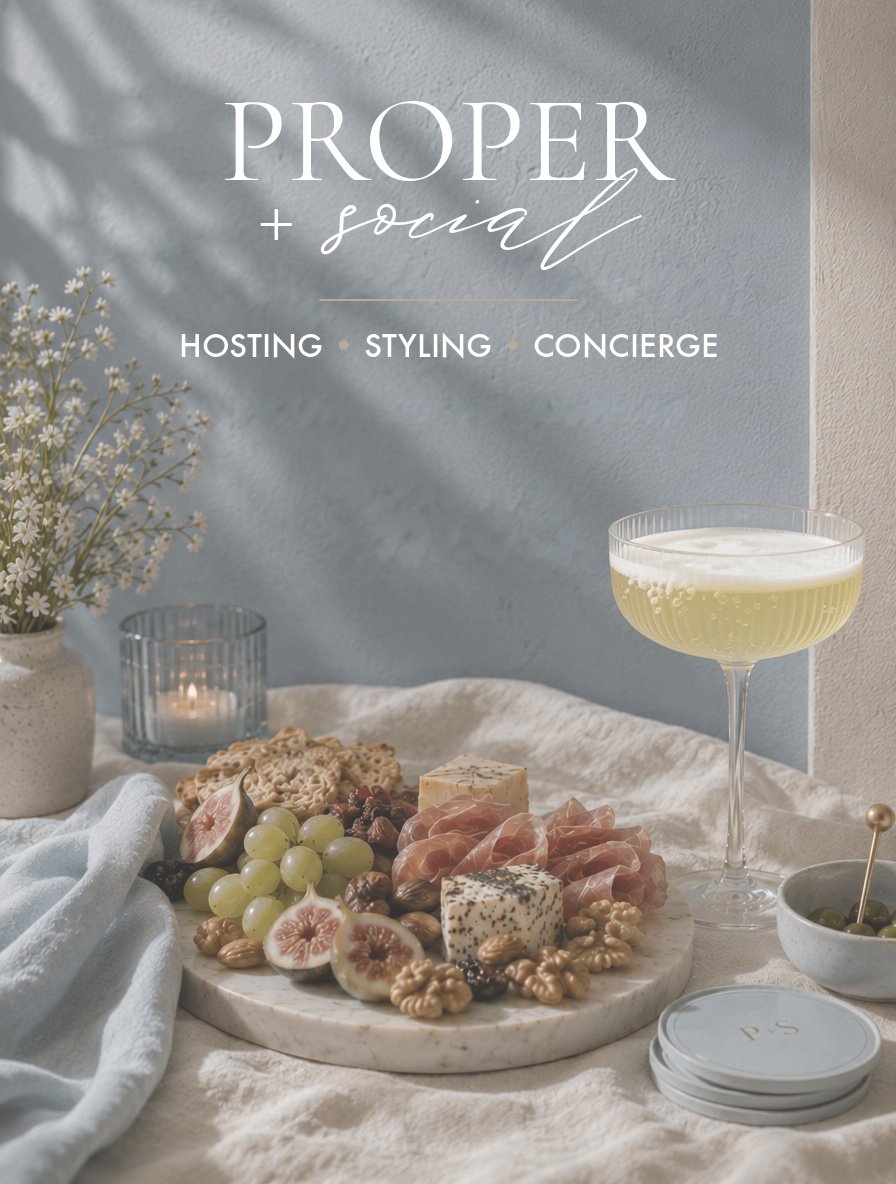Decorative table setting with cheese, grapes, crackers, and cured meats on a marble platter, a glass of champagne, a lit candle, a bowl of green olives, and a flower arrangement, with a sign that reads 'Proper + Social' indicating hosting, styling, and concierge services.