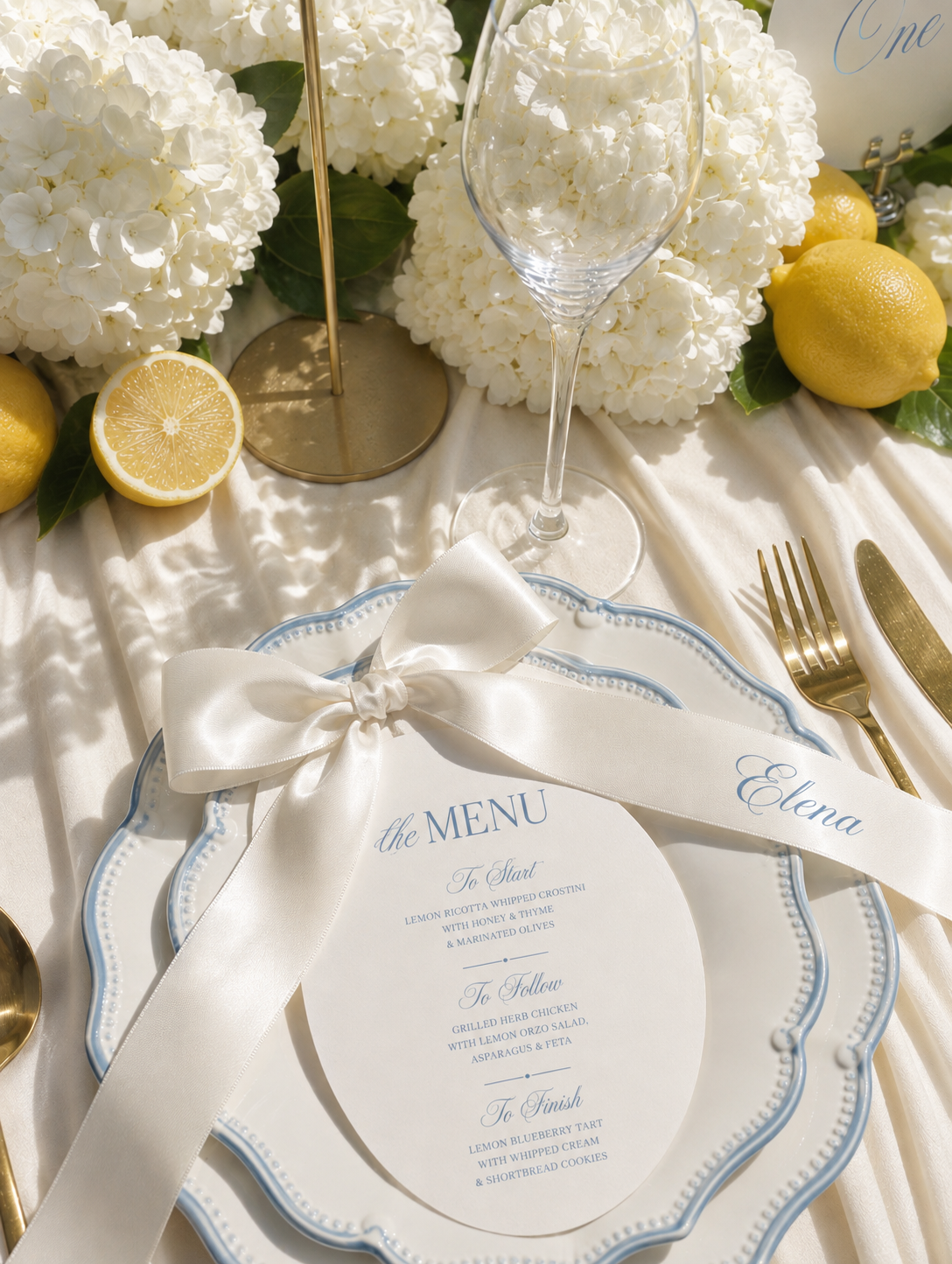 Elegant table setting with a menu, white hydrangea flowers, lemons, a wine glass, and gold cutlery, decorated for a special occasion.