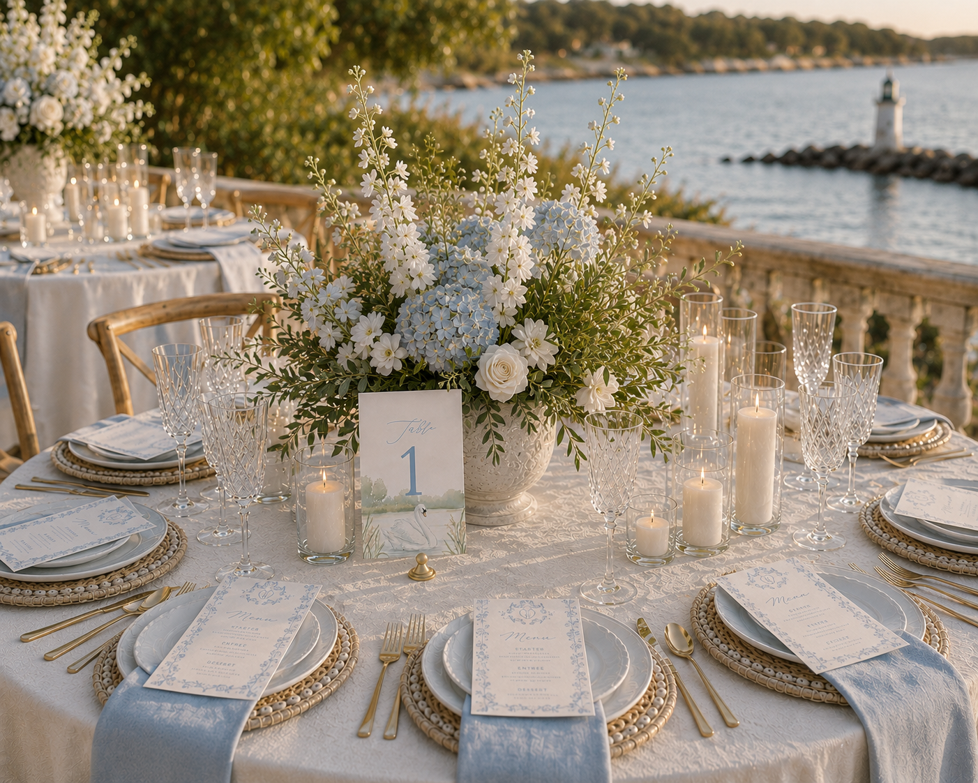 A beautifully set outdoor banquet table overlooking a body of water with a lighthouse in the background. The table features a large floral centerpiece with white and blue flowers, surrounded by candles in glass holders. Elegant place settings include white plates, gold flatware, crystal glasses, and printed menus. A table number card with a swan illustration is also displayed.