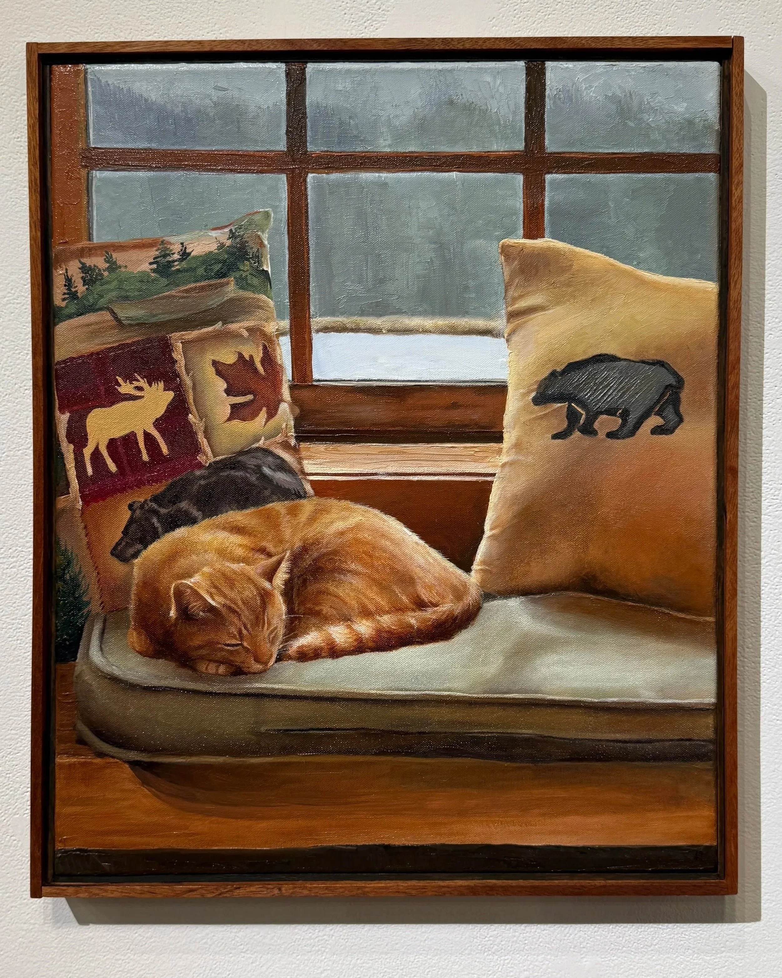 Painting of a sleeping orange tabby cat on a windowsill with woodland-themed pillows, a large beige pillow with a bear silhouette, and a view of a snow-covered landscape outside the window.