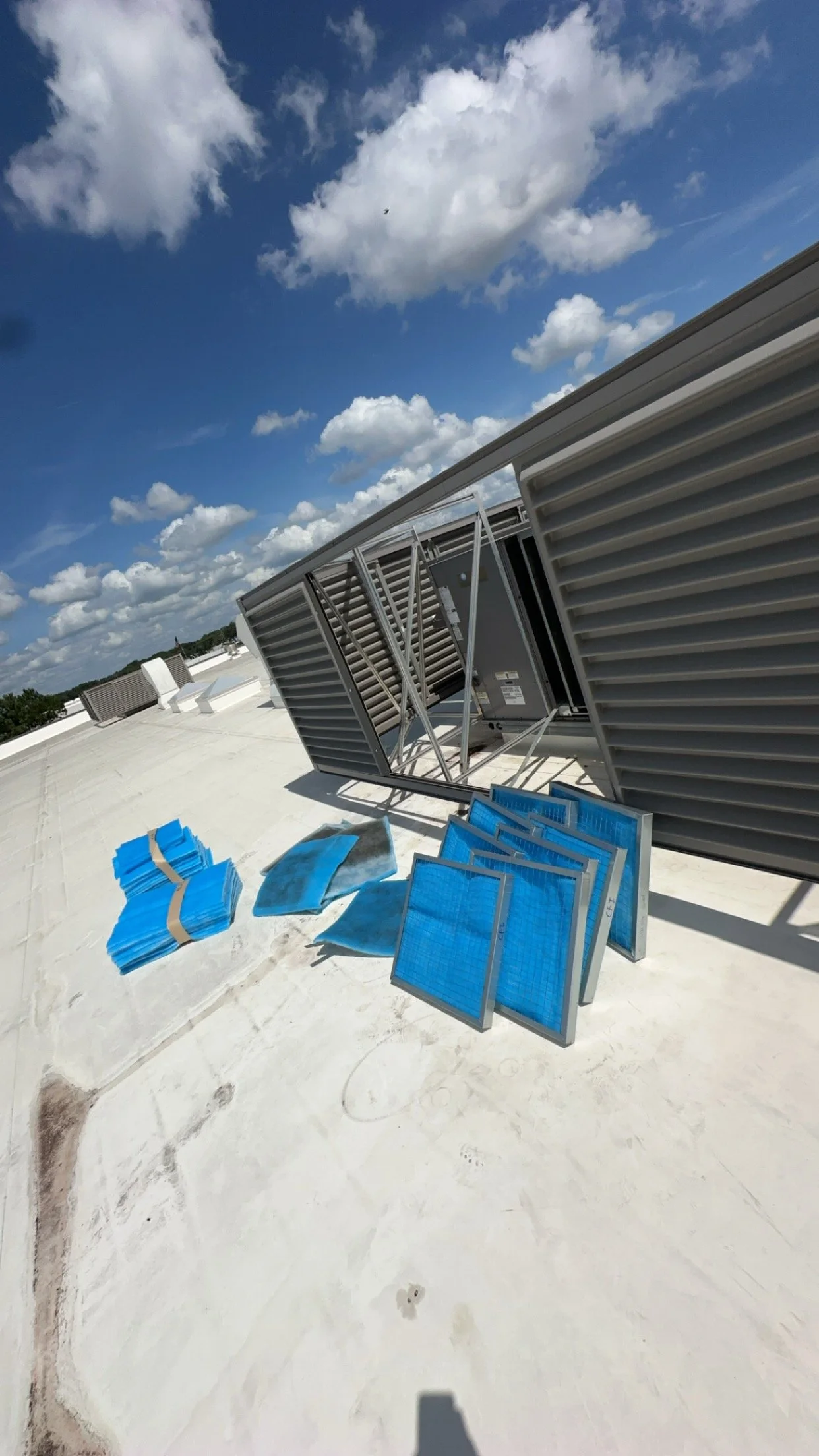 Air conditioning unit components on a rooftop, including blue filters, metal frames, and vents, with a partly cloudy sky in the background.