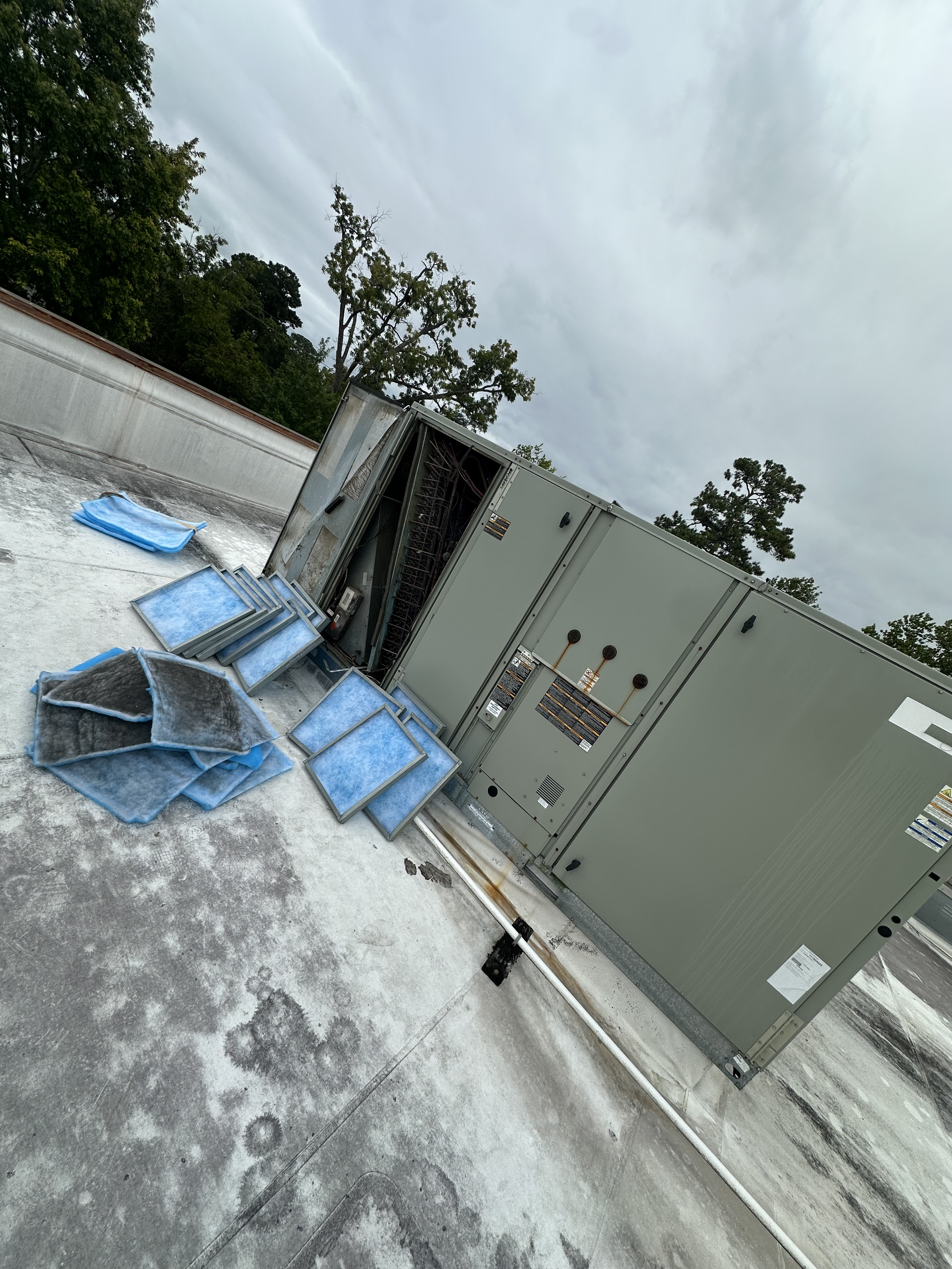 Air conditioning unit on a flat rooftop with removed filter panels and tools nearby, cloudy sky in the background.