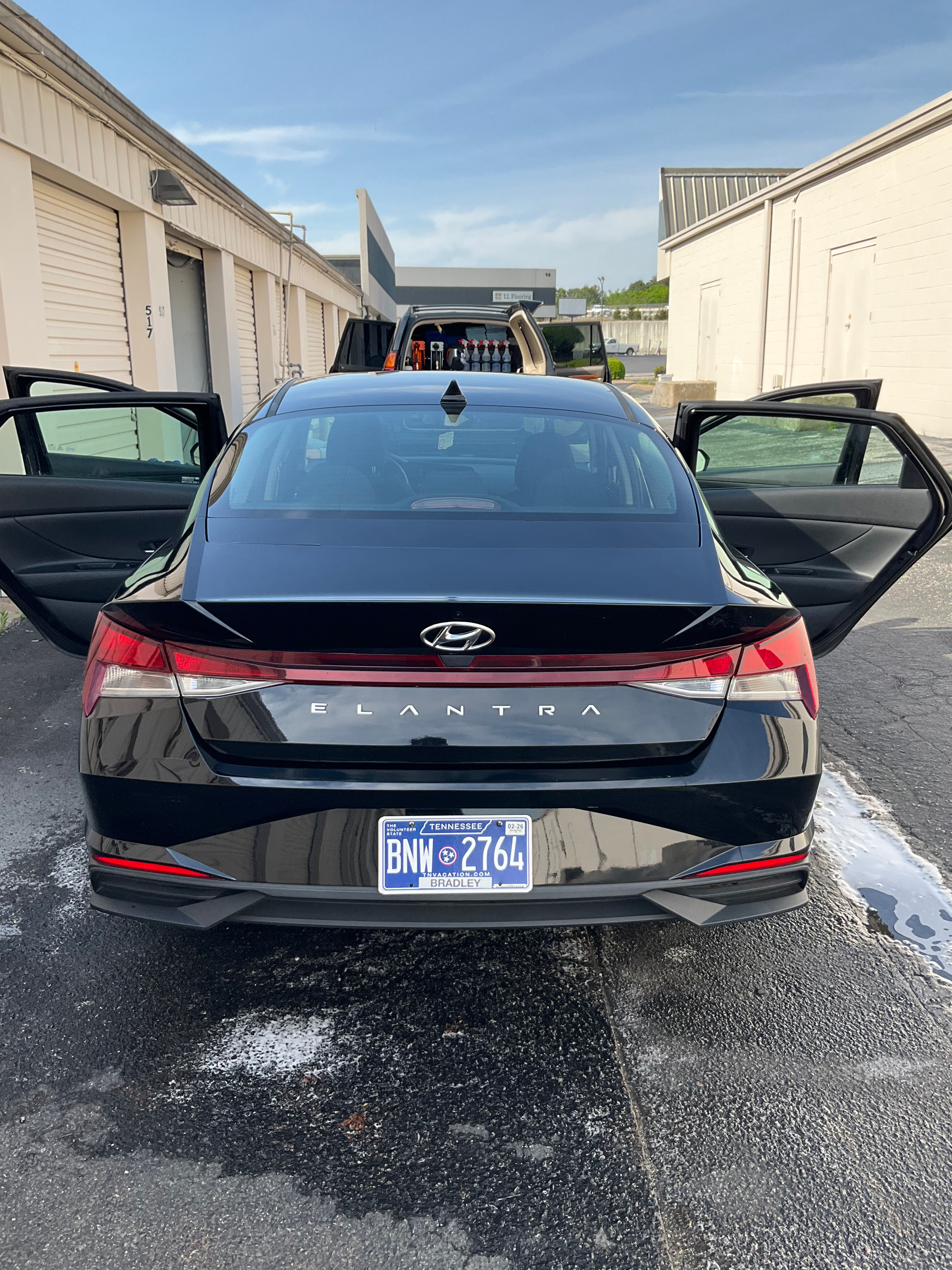 Rear view of a black Hyundai Elantra with open doors, parked near storage units. Tennessee license plate visible. A van with supplies in the background. Clear sky above.