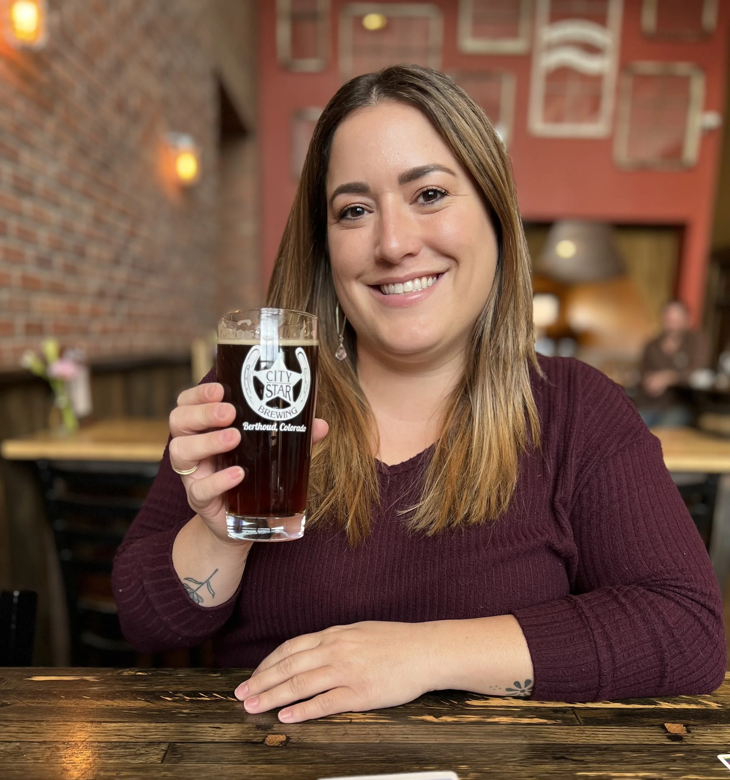 Emily smiling at the camera, sitting at a table, and holding a glass of dark beer.