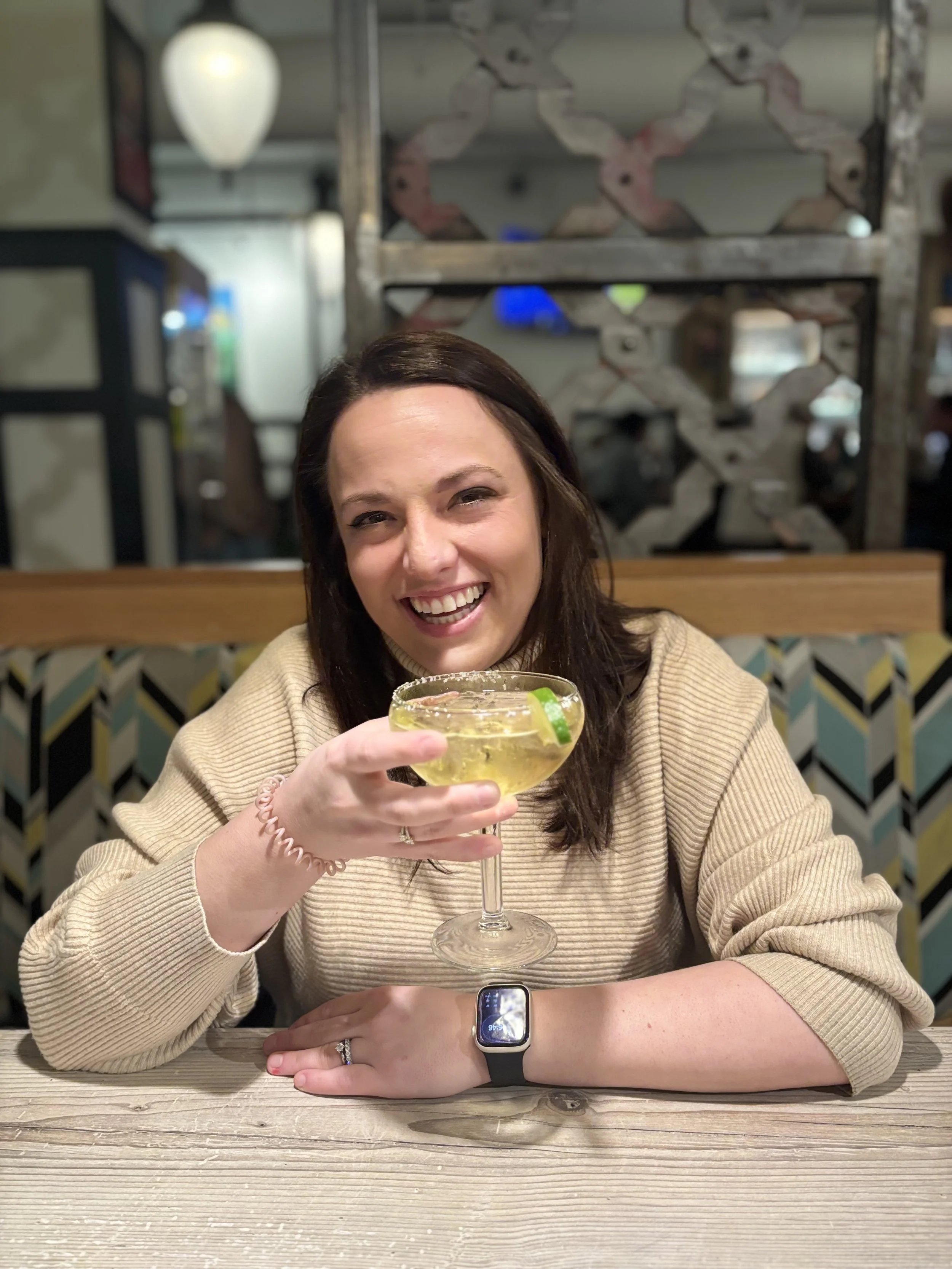 Tori smiling at the camera, sitting at a table, and holding a margarita.
