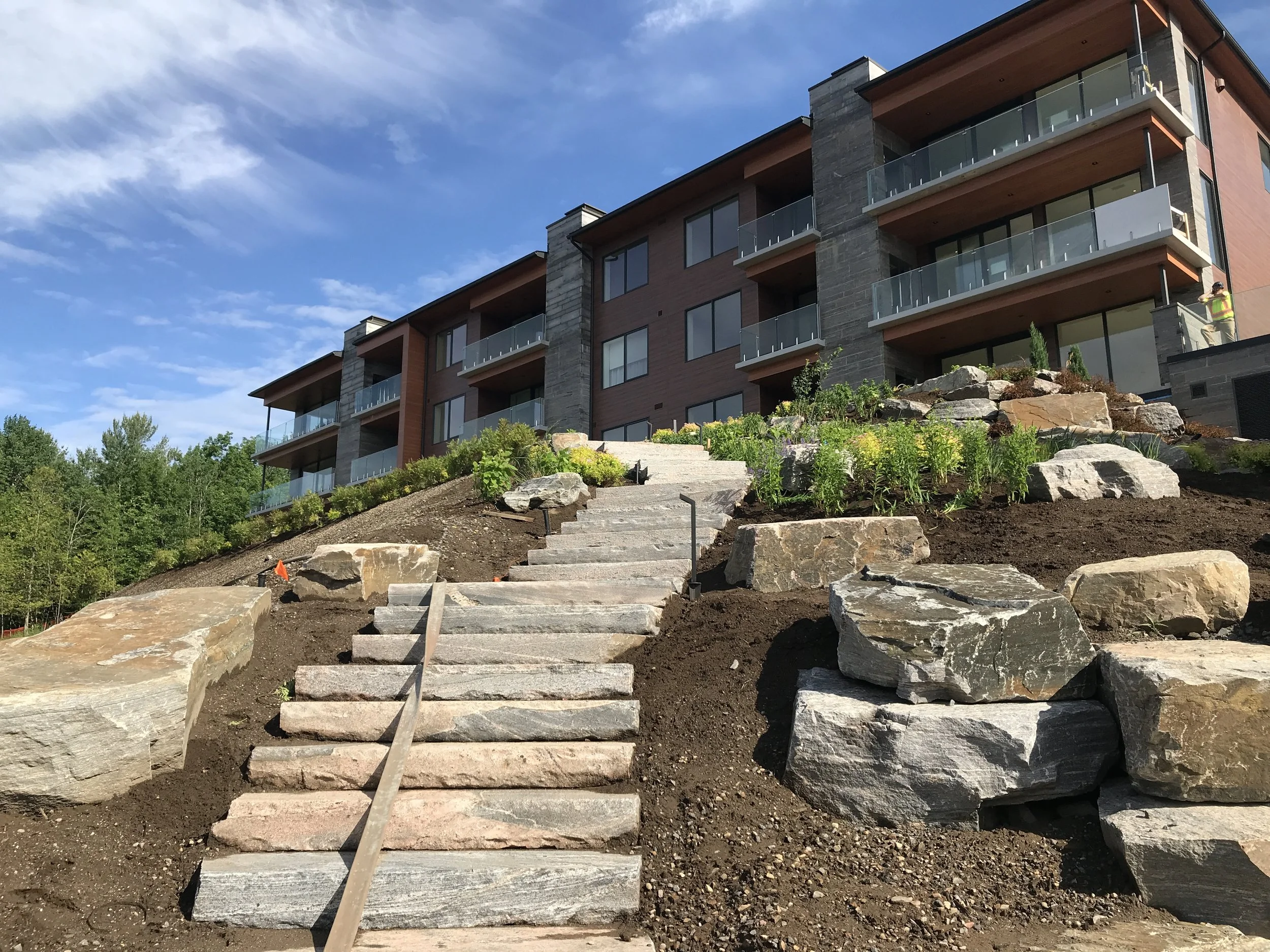 A newly landscaped outdoor area with stone steps and large rocks leading up to a modern multi-story apartment building with balconies, surrounded by greenery under a partly cloudy sky.