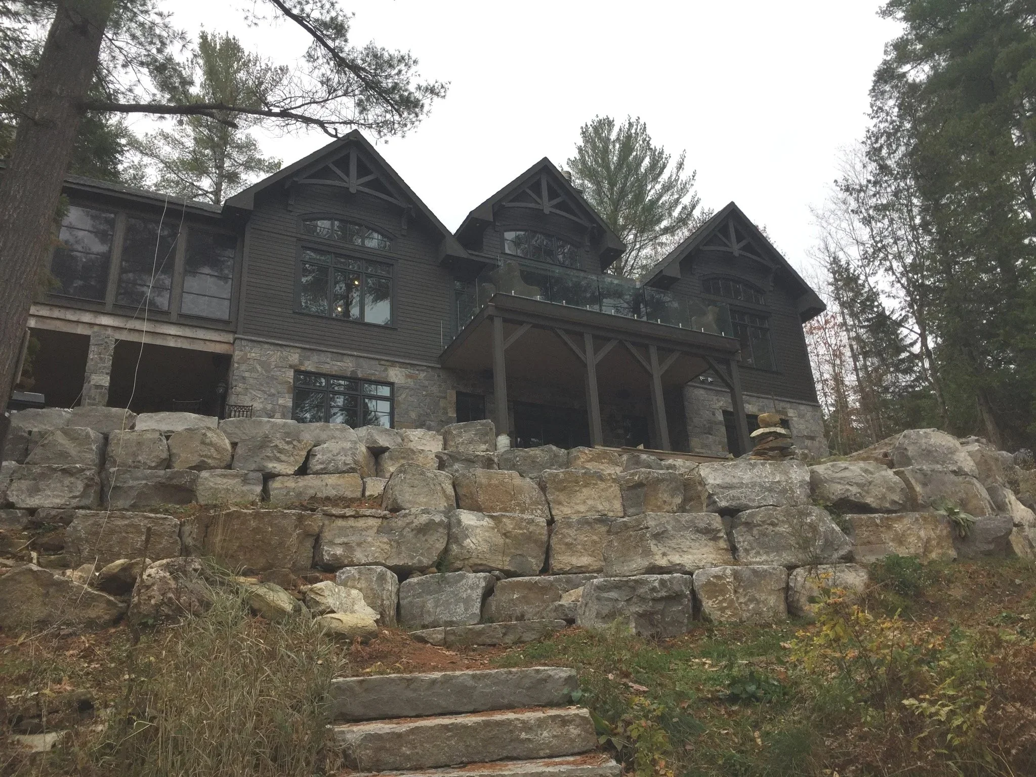 A large, multi-story house built on a hill with a rock retaining wall and stone steps leading up to it, surrounded by trees.