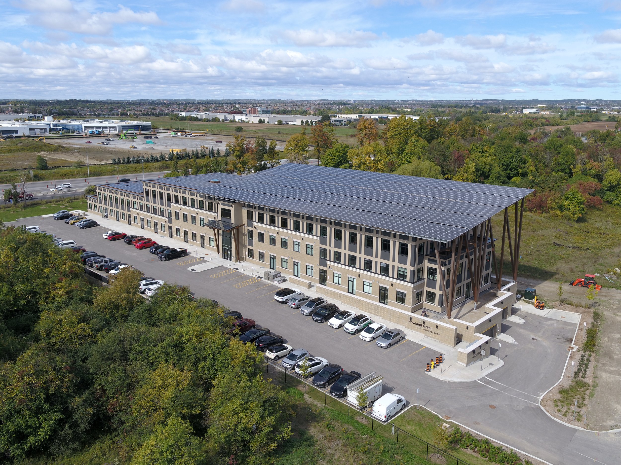 An aerial view of a modern three-story building with solar panels on the roof, surrounded by a parking lot with several cars, trees, and a distant industrial area under a partly cloudy sky.