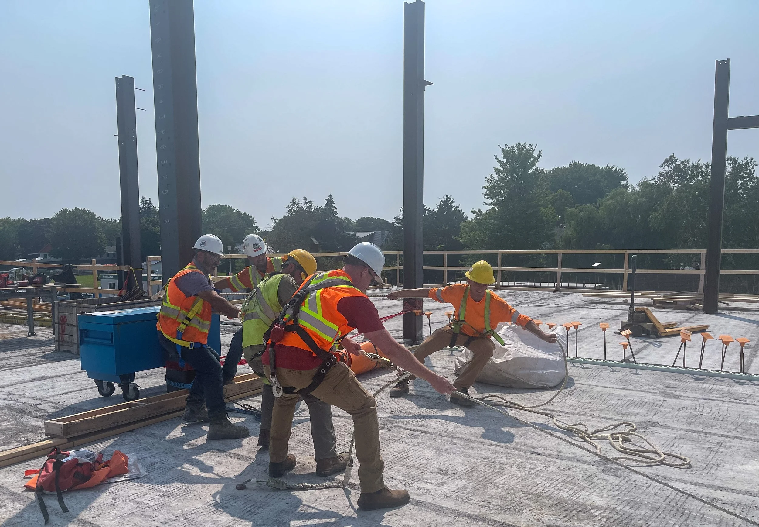 Construction workers on a building site, wearing safety helmets and vests, working with ropes and tools on a rooftop under construction.