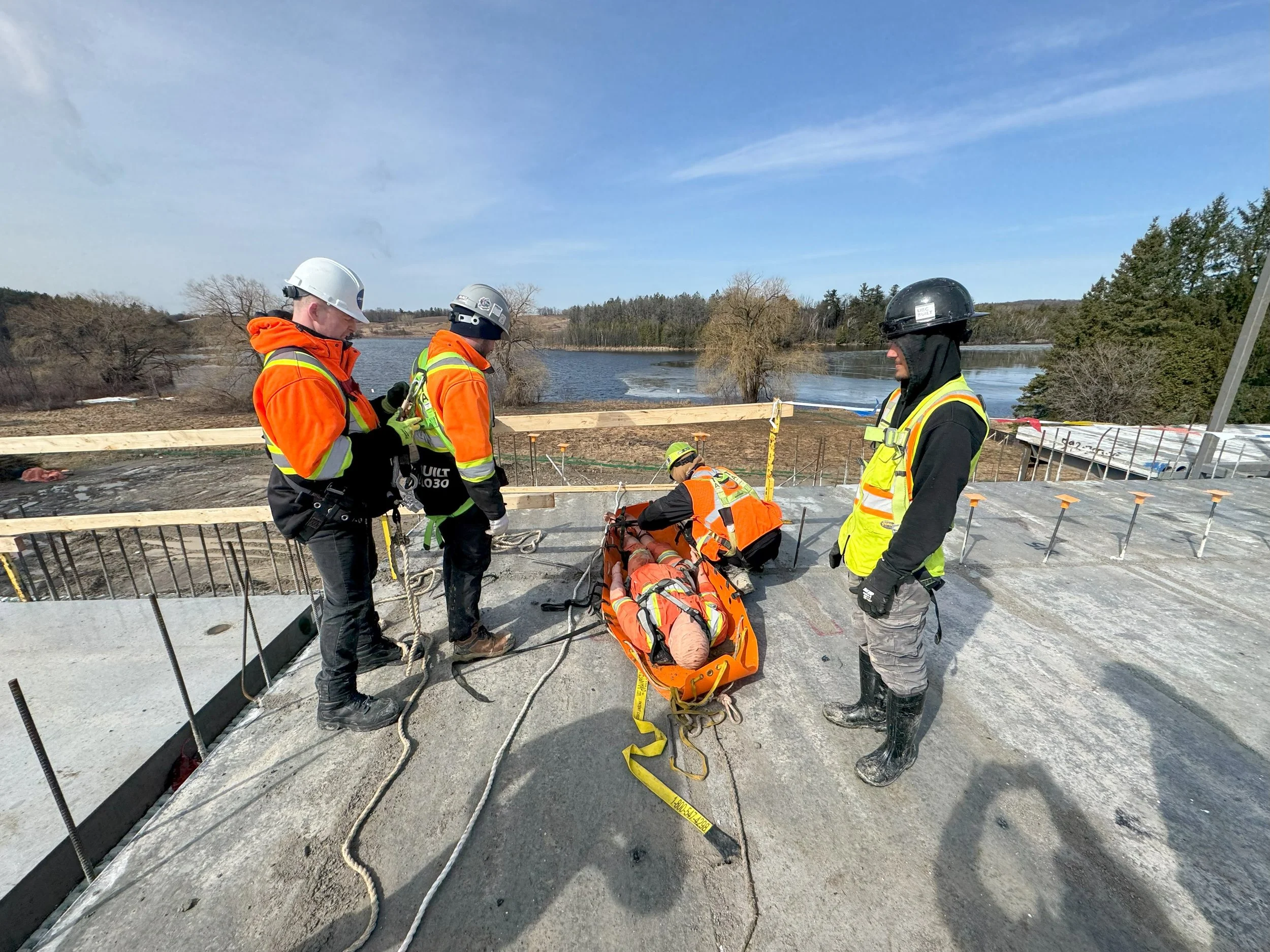 Construction workers on a bridge, wearing safety gear, preparing to lift a person on a stretcher with ropes, overlooking a river with a partly cloudy sky.