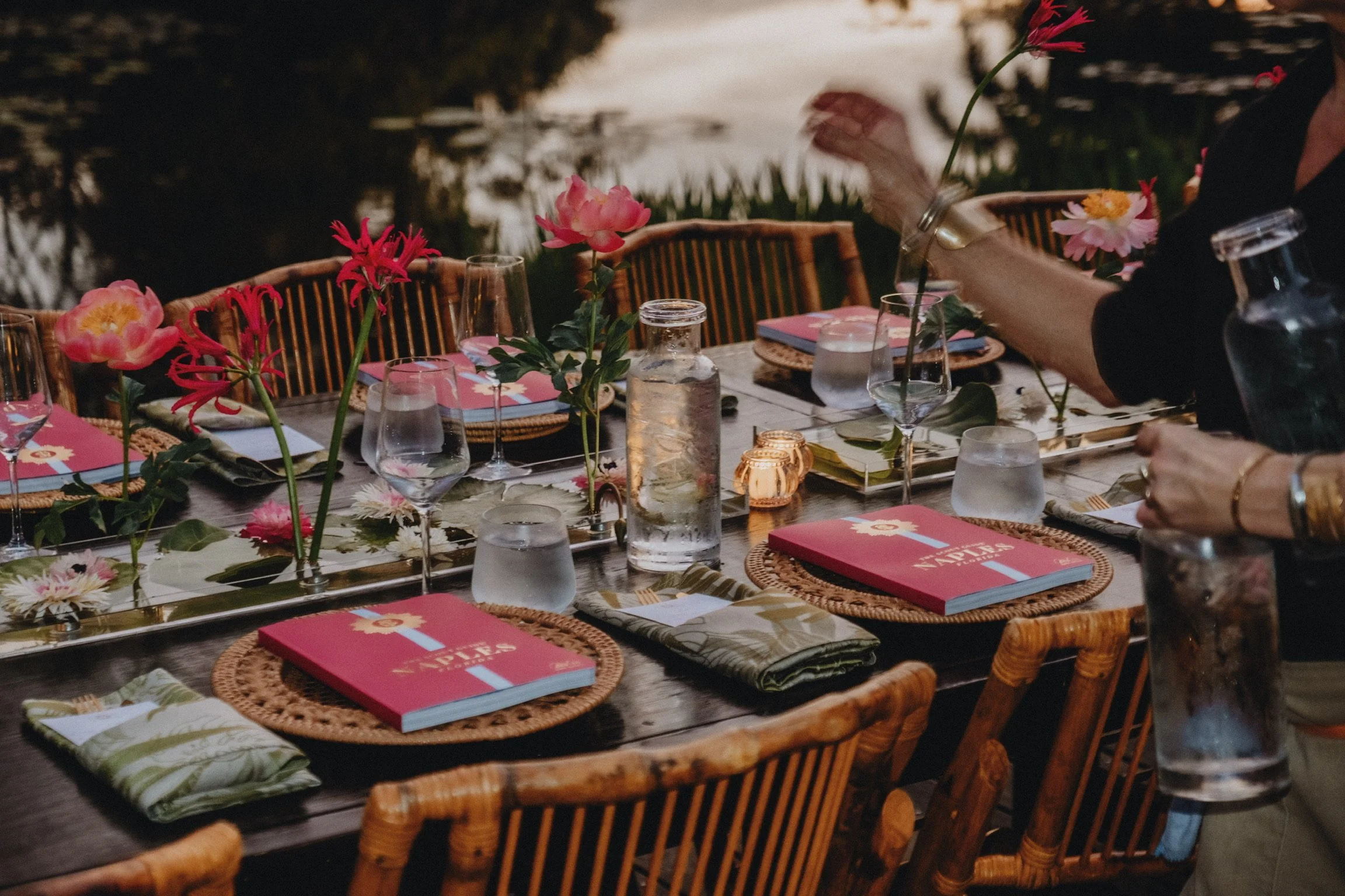 A dinner table set outdoors with pink flowers and candles, featuring place settings with glassware, cloth napkins, and pink menus, near a body of water during sunset.