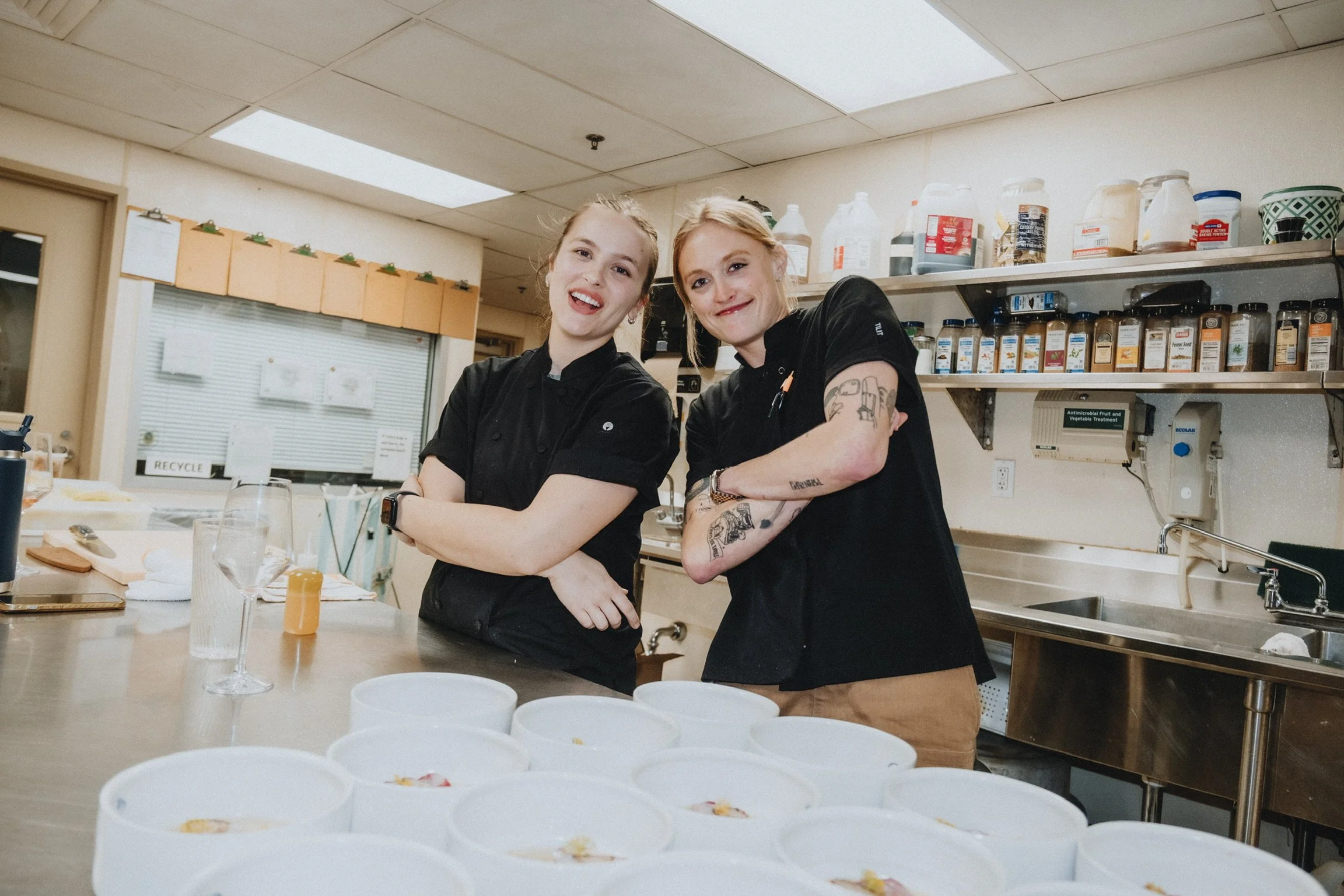 Two women stand behind a kitchen counter with bowls of food in front of them, smiling and posing for the camera in a busy kitchen or restaurant setting.