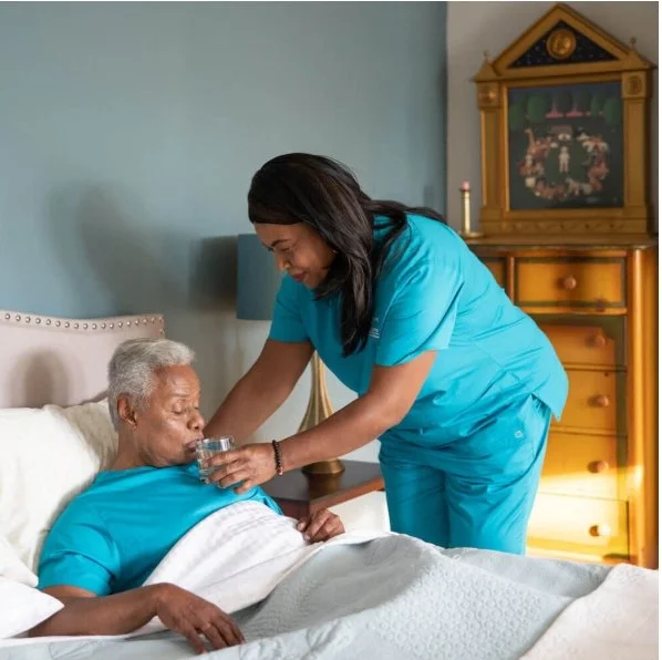 A caregiver in blue scrubs offers a glass of water to an elderly woman lying in bed, in a cozy bedroom with light blue walls, a wooden dresser, and a framed picture on top.