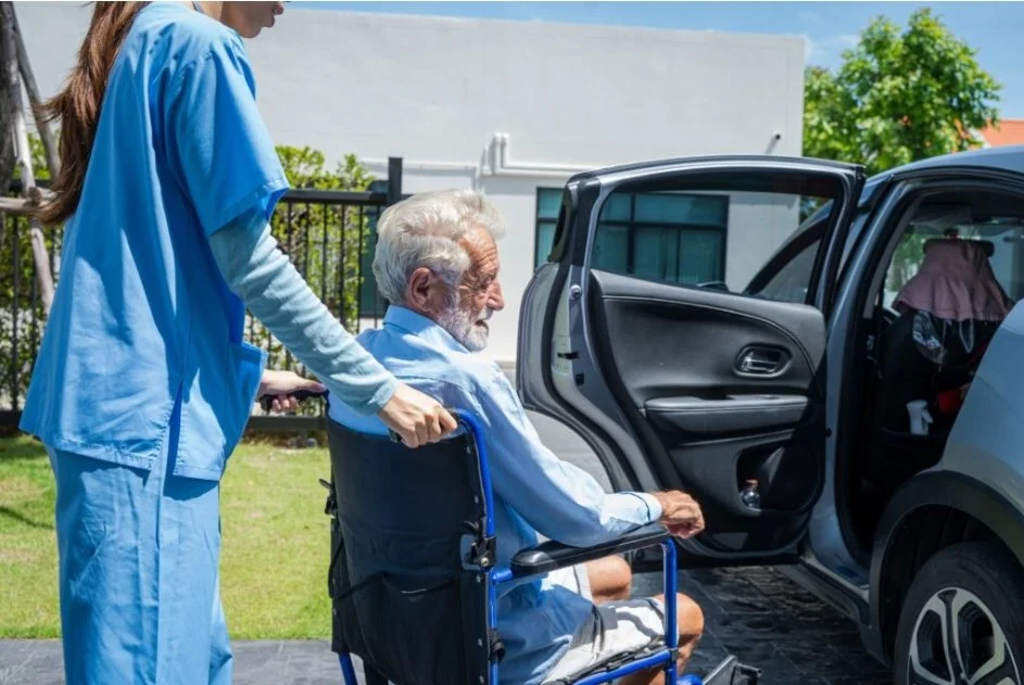 Nurse pushing an elderly man in a wheelchair towards a car for transportation.