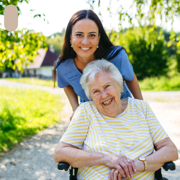 A young woman in blue scrubs smiling and leaning over an elderly woman in a wheelchair outdoors.