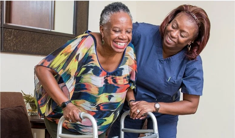 A woman using a walker is supported by a healthcare professional who is assisting her, both smiling in a healthcare setting.