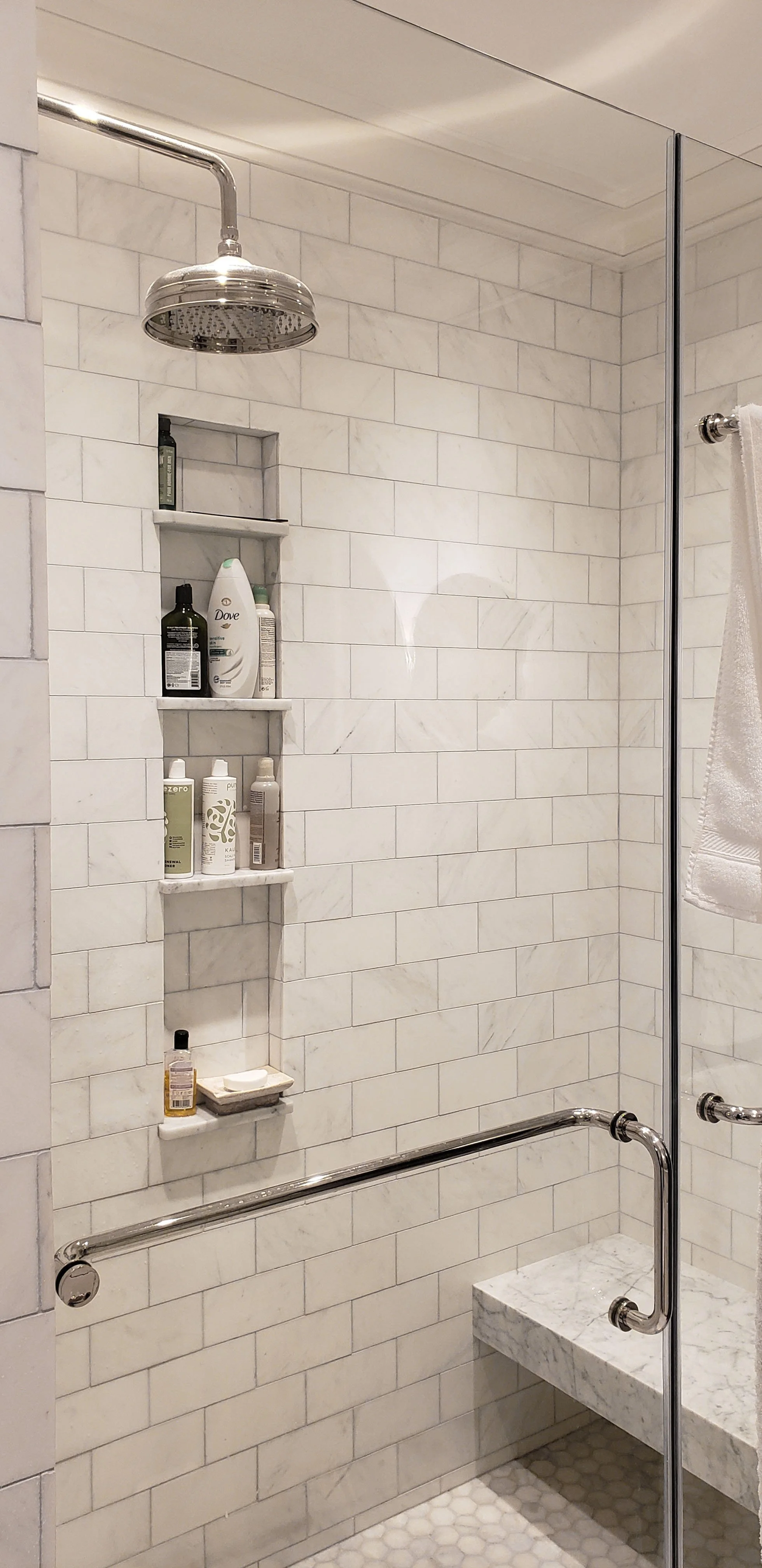 Shower area with white subway tiles, a built-in niche with personal care products, a rain shower head, and a marble seating bench with a white towel hanging.