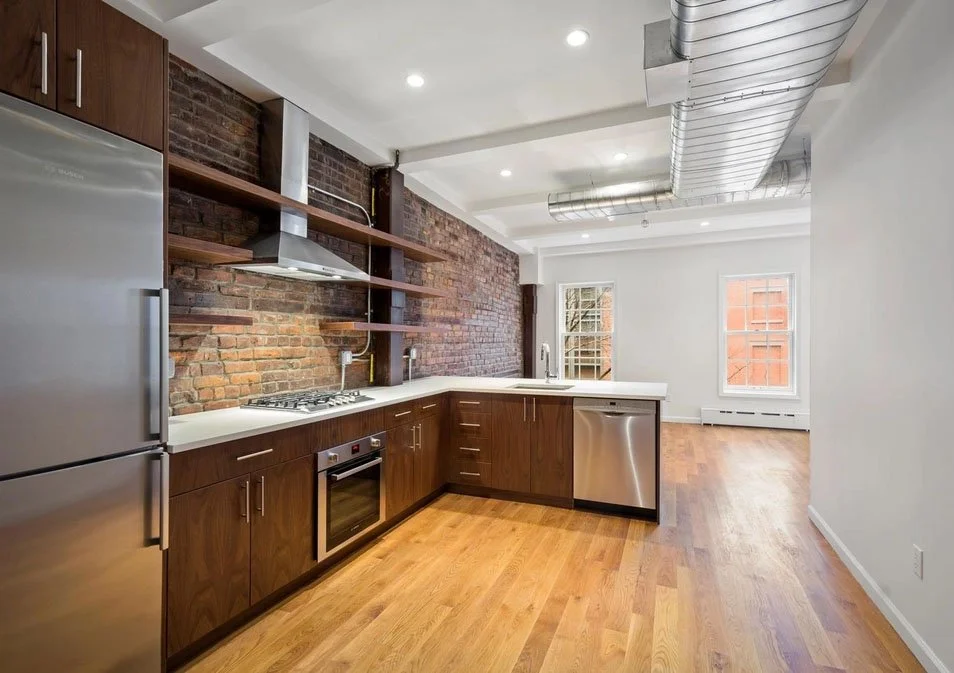 Modern kitchen with exposed brick wall, stainless steel appliances, cherry wood cabinets, open shelving, and large windows in an industrial style space.