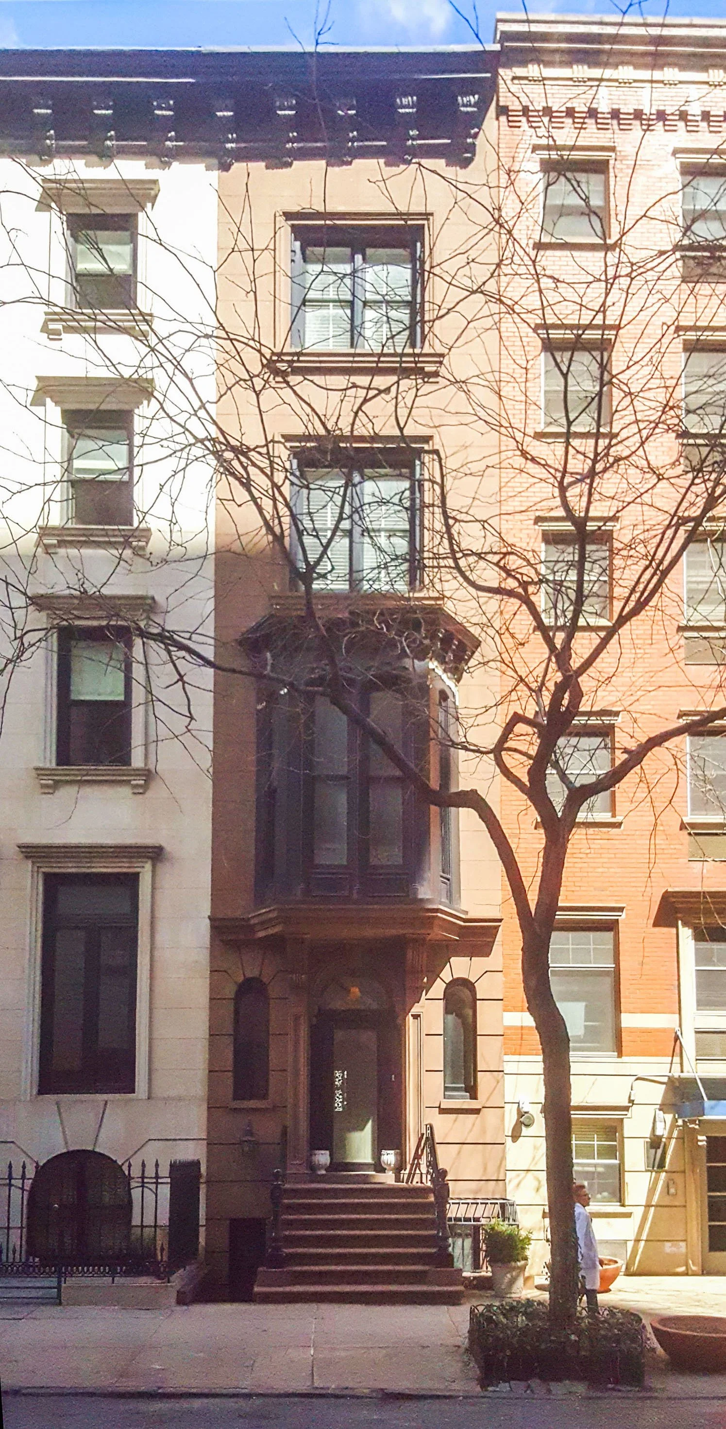 Facade view of a brownstone townhouse with a bay window and steps leading to the entrance, flanked by a tree and a person standing on the sidewalk.
