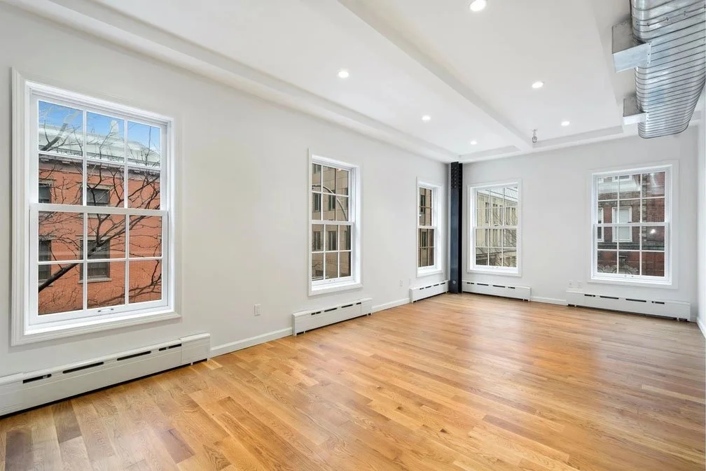Empty room with white walls, hardwood floors, several windows, ceiling lights, and exposed ductwork.