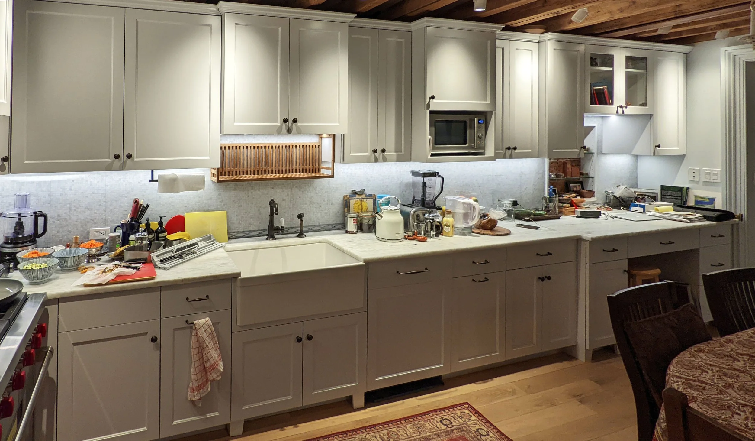 A cluttered kitchen counter with various appliances, utensils, and food items, with white cabinets and a wooden ceiling.