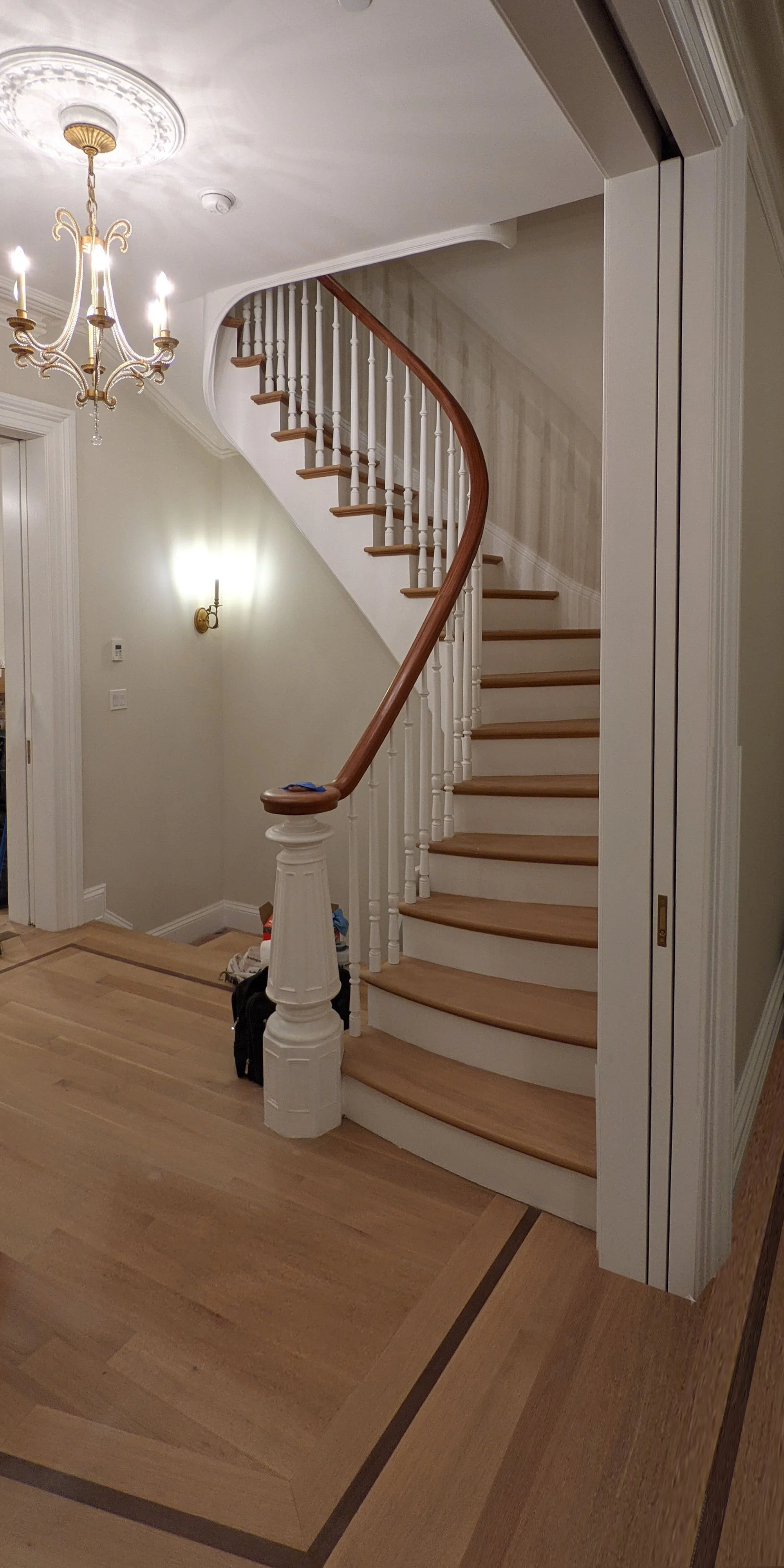 Interior view of a staircase with wooden steps and white balusters, adjacent walls painted in light colors, a chandelier hanging from the ceiling, and a wall sconce light fixture.