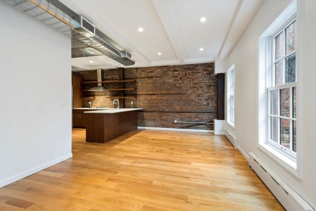 Empty apartment with hardwood floors, large windows, white walls, and an exposed brick wall in the kitchen area.
