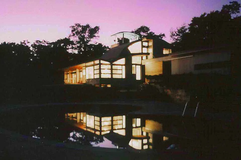 A house illuminated from within at dusk, with trees in the background and their reflection visible on a body of water in the foreground.