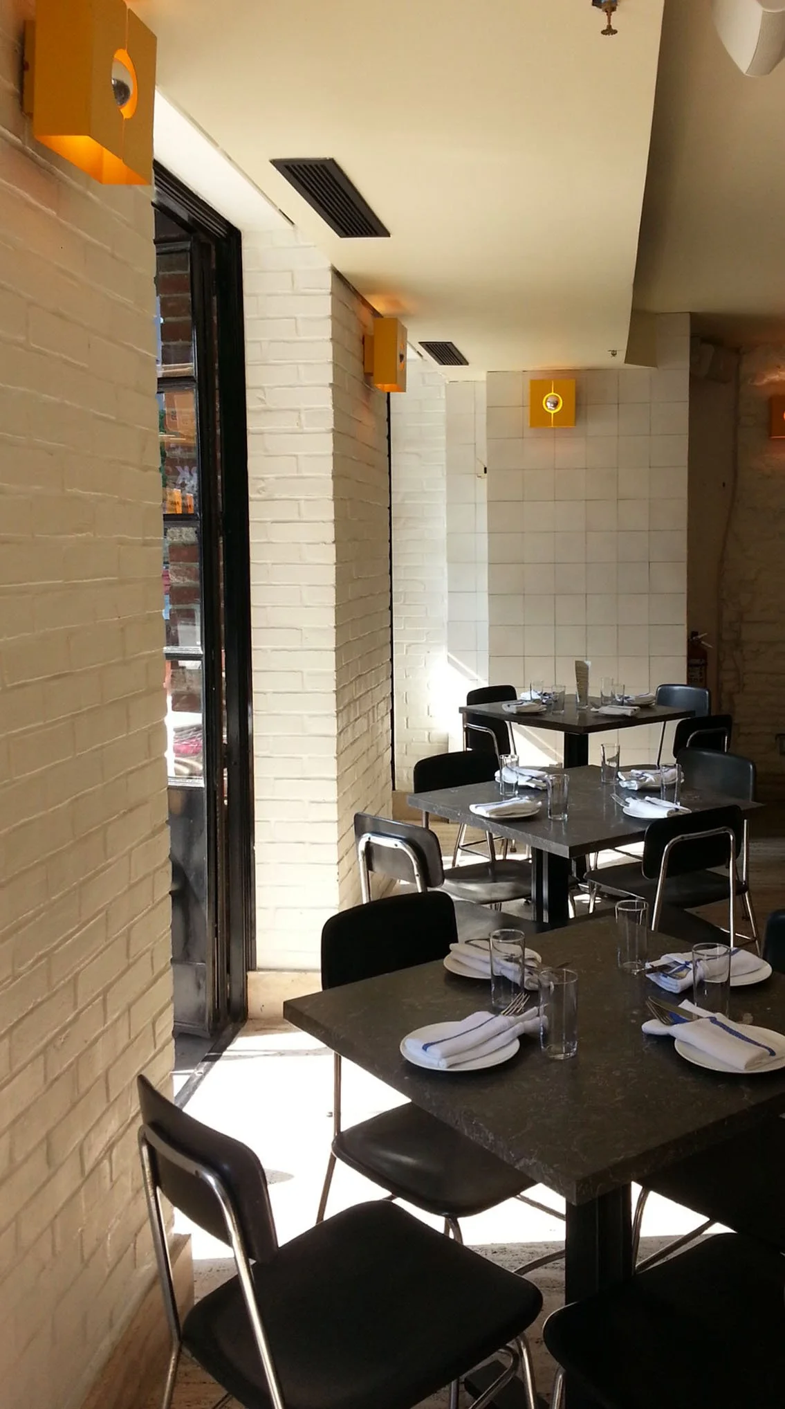 Inside a restaurant with tables set with napkins, glasses, and utensils, featuring yellow wall-mounted lights and a partially open door leading outside.