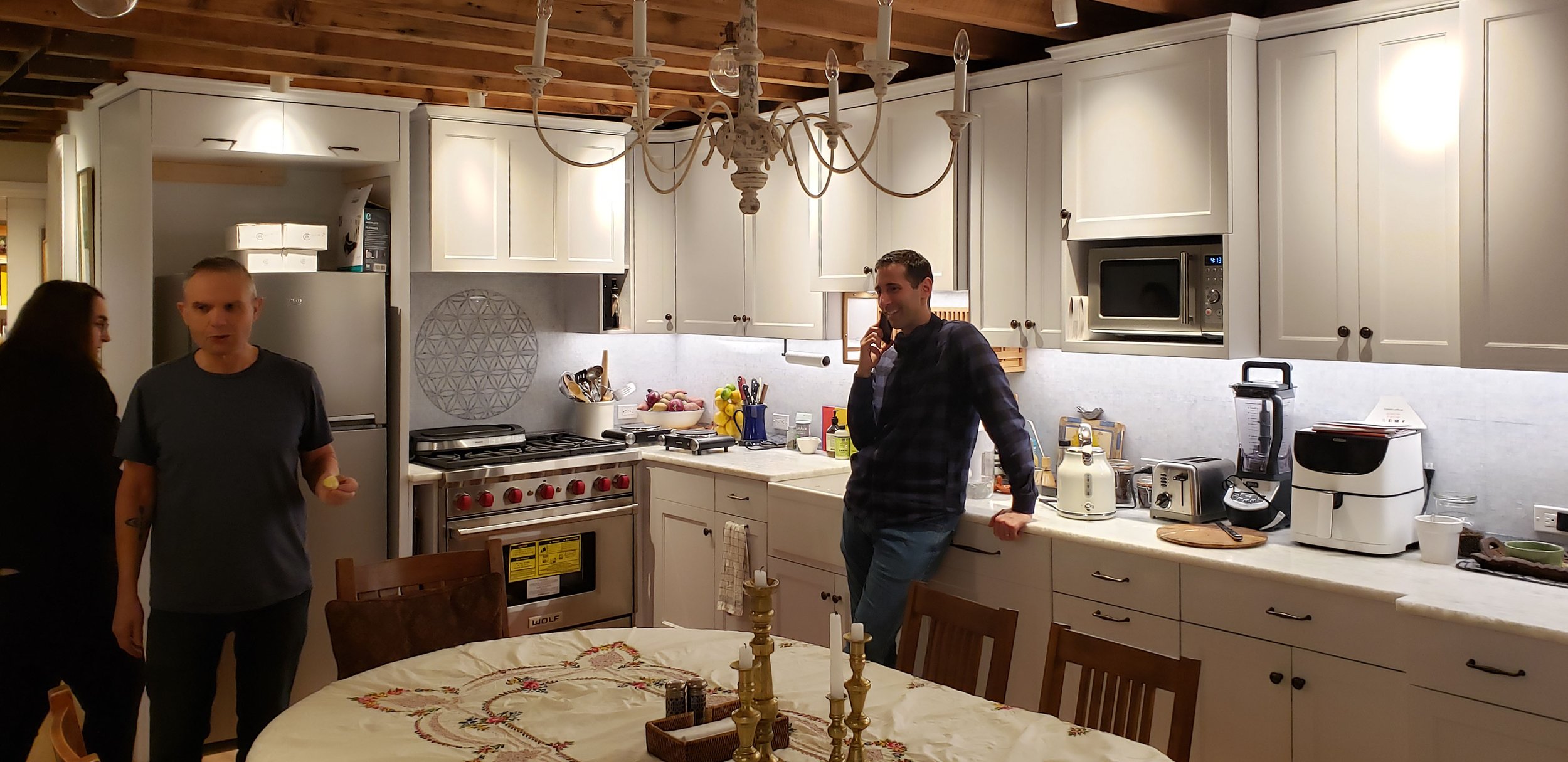 A cozy kitchen scene with three people, a woman and two men, engaged in conversation. The kitchen has white cabinets, a chandelier, a stove, microwave, and various appliances on the counter. A large dining table with a decorative centerpiece is in the foreground.