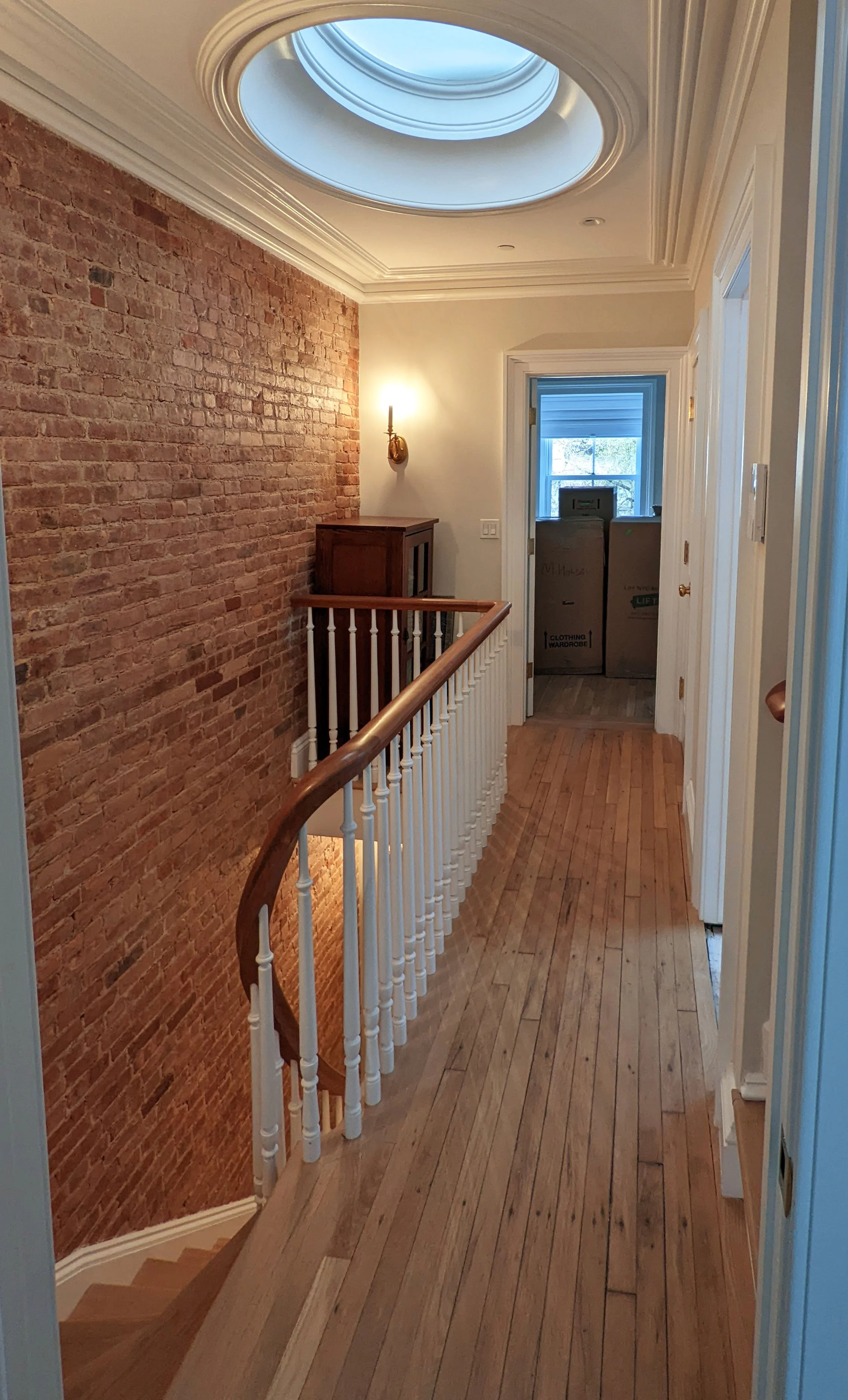 Hallway with wooden flooring and a brick wall on the left. A staircase is visible at the bottom left, descending downward. A small wooden cabinet and a wall-mounted light fixture are on the brick wall. On the right side, there are several doors, one slightly open, revealing boxes inside a room with a window. The ceiling has decorative molding and an oval skylight allowing natural light in.