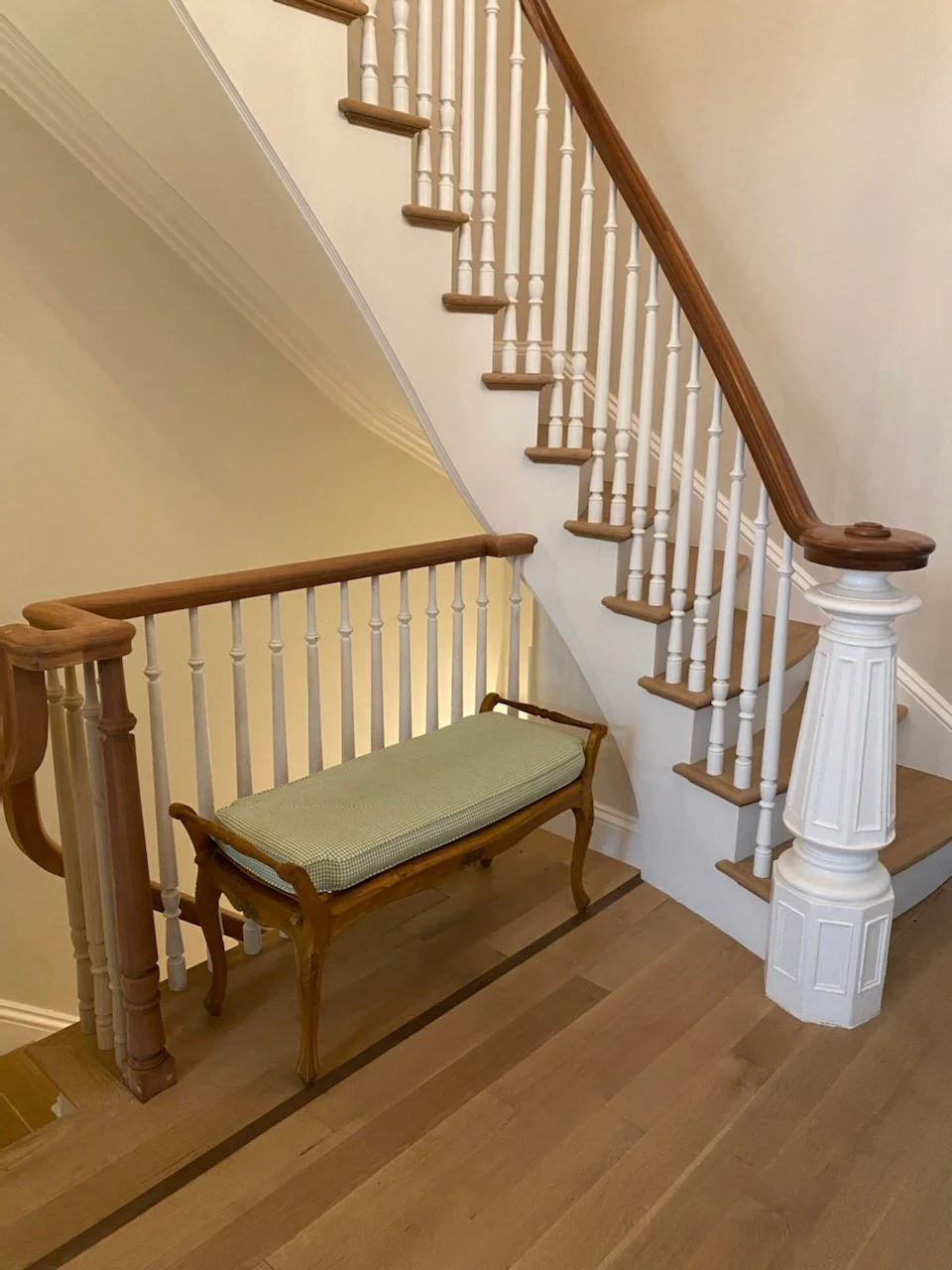 Wooden staircase with white spindles and brown handrail, a vintage wooden bench with a green cushion, hardwood flooring, cream-colored walls, and decorative white molding.