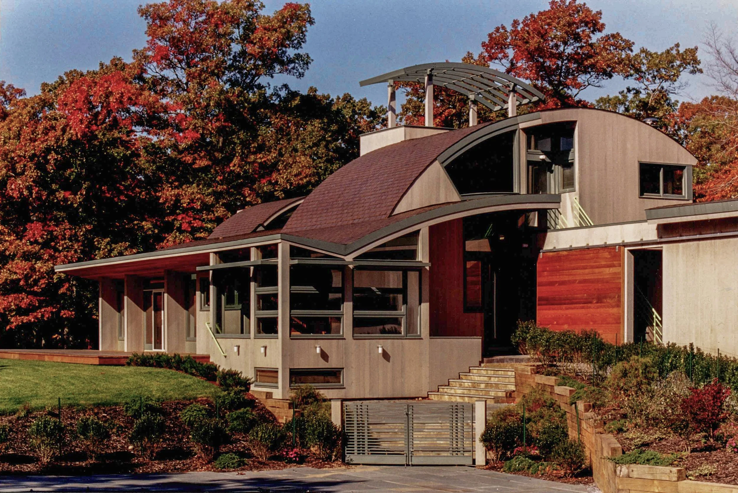 Modern architectural house with curved roof sections, surrounded by autumn trees with colorful foliage, a manicured lawn, and a gated entrance in the foreground.
