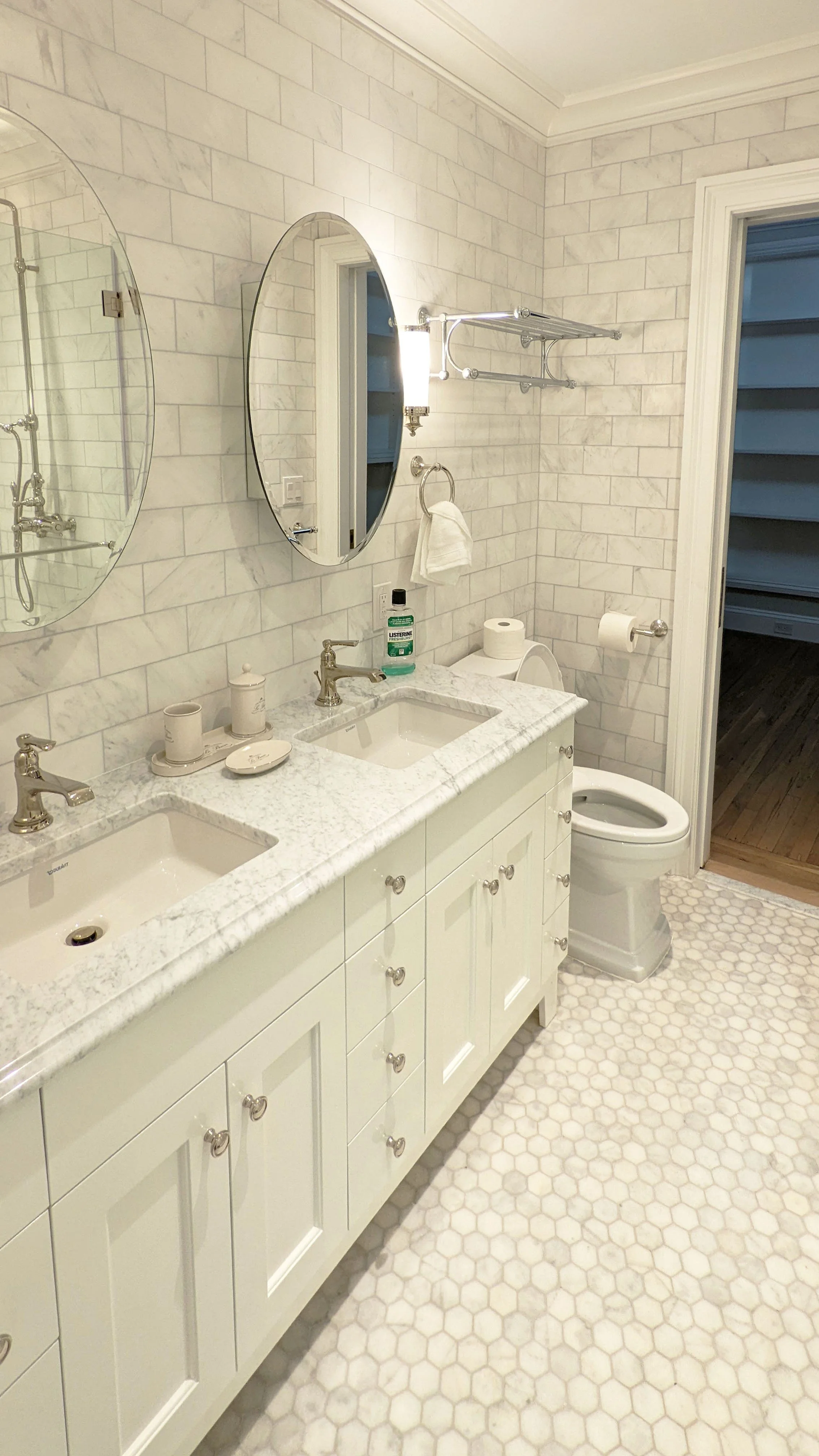 Bathroom with white marble countertop double sinks, oval mirrors, wall-mounted light fixture, toilet, and hexagonal tile flooring.