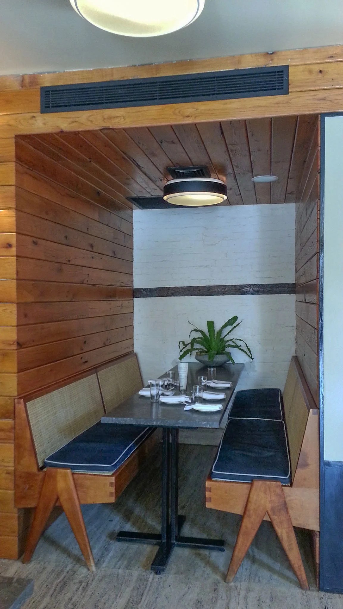 A private dining nook with a wooden benches, a table set with glasses and napkins, and a potted plant on the white wall in the background. The room has wood-paneled walls and a ceiling with a round black light fixture.