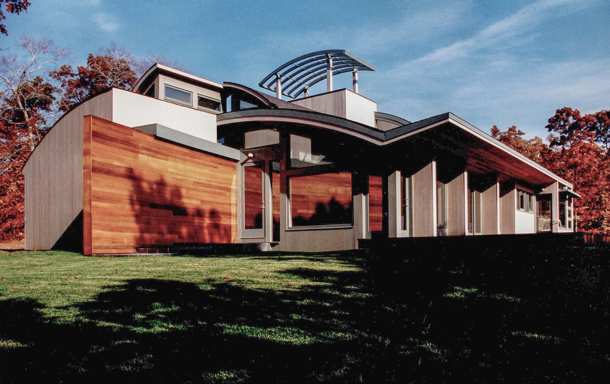 A modern multi-story house with a mix of wood and concrete exterior, large windows, and an outdoor covered balcony, set against a backdrop of autumn trees and a clear blue sky.