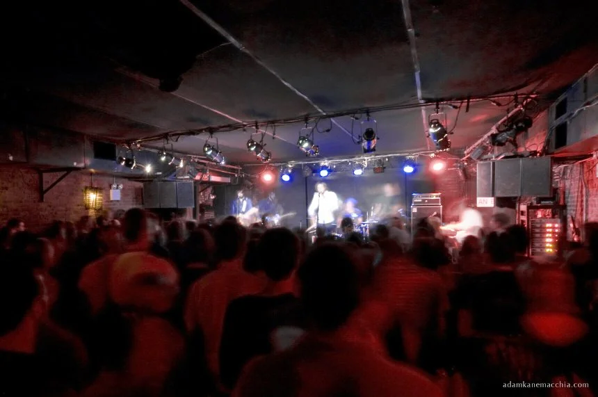 Crowd watching a live band performance on stage at a small indoor concert venue with colorful stage lighting.