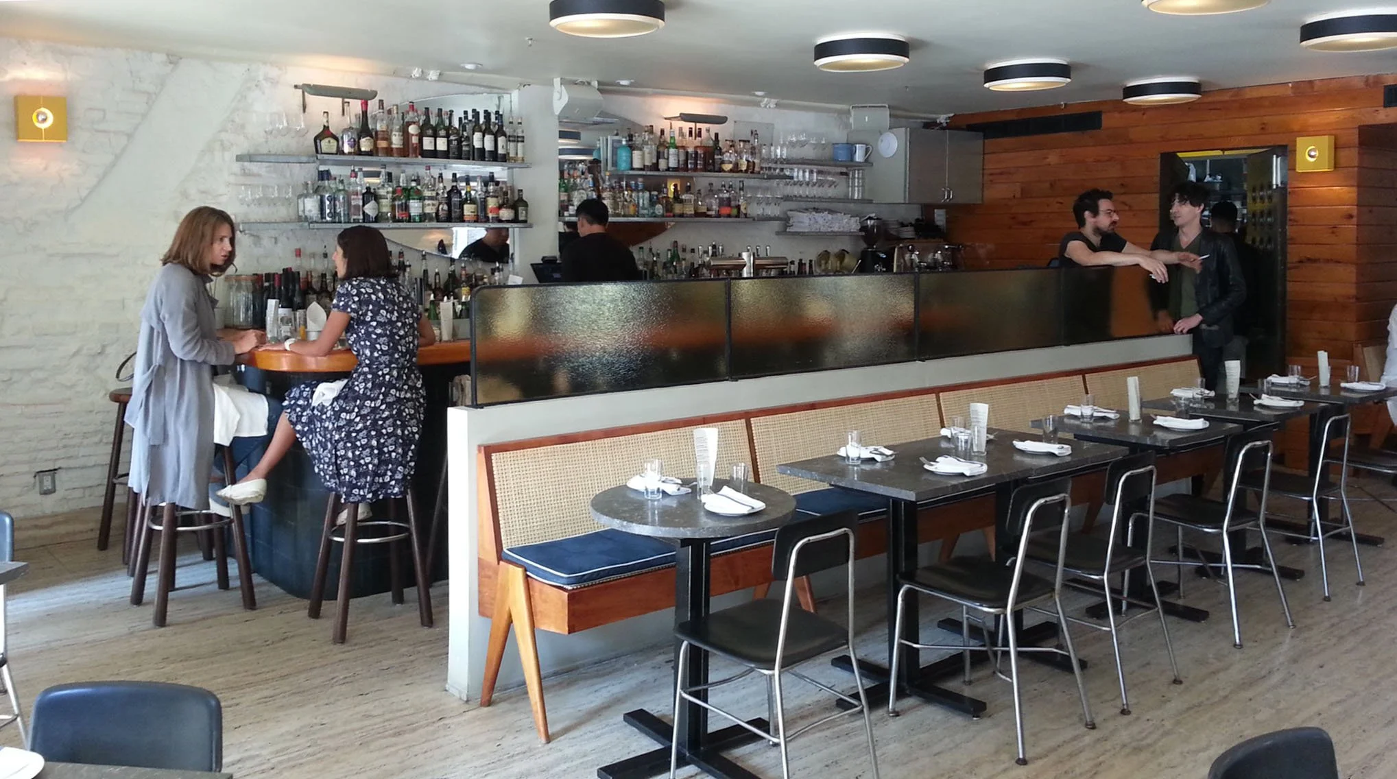 Interior of a restaurant with tables, chairs, a bar with bottles, and three people talking near the bar.