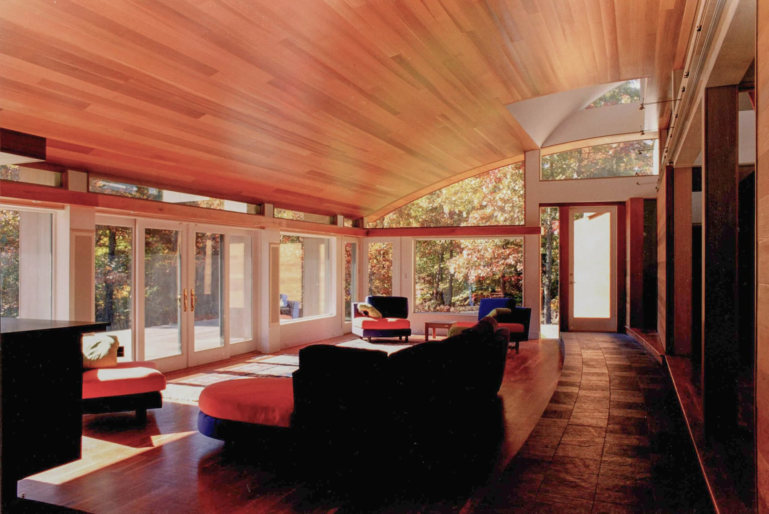 Sunlit interior of a modern living room with large windows, wooden floors, and contemporary furniture including black and red chairs, and a stone tile floor.