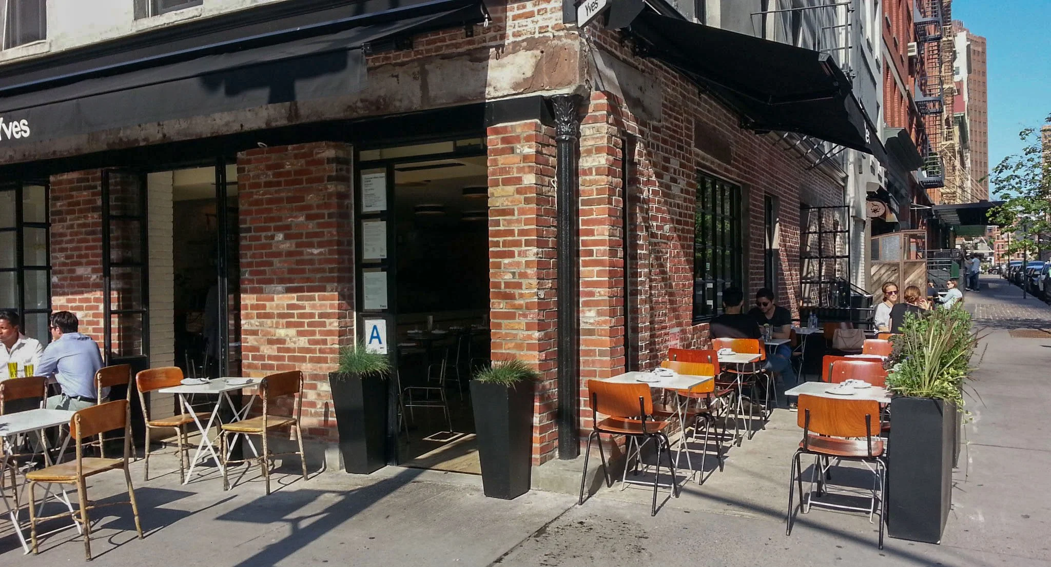 Outdoor sidewalk cafe with people dining in front of a brick building during daytime.