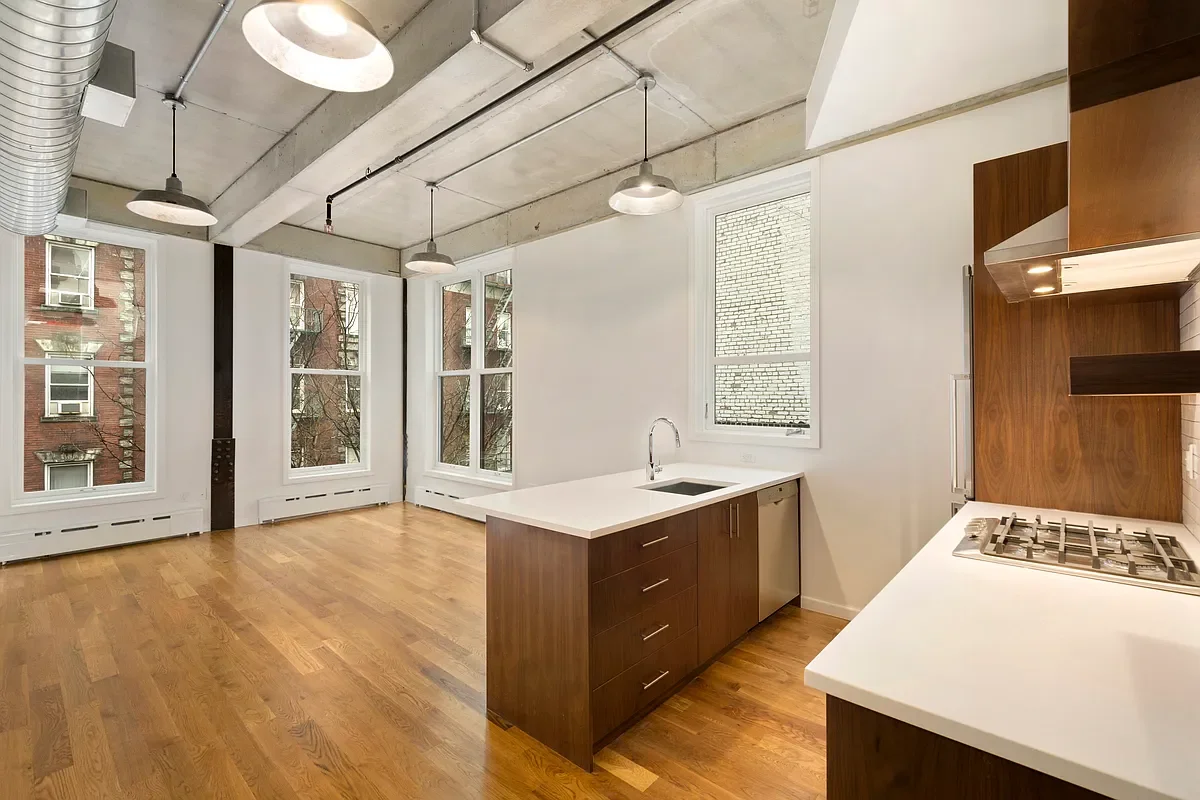 Open-concept modern kitchen with wood floors, white countertops, and exposed concrete ceiling. Large windows allow natural light, with three hanging industrial-style pendant lights. Kitchen features a central island with a sink and a stove on the adjacent counter.