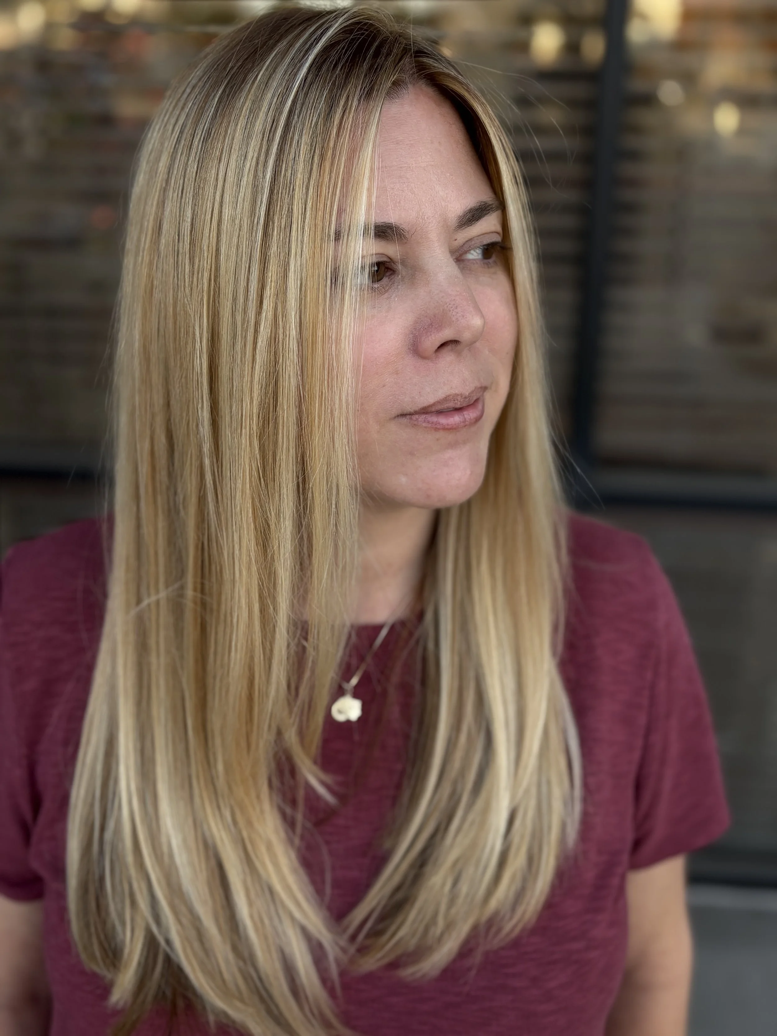 A woman with long blonde hair in a maroon shirt, looking to the side, with a blurred background.