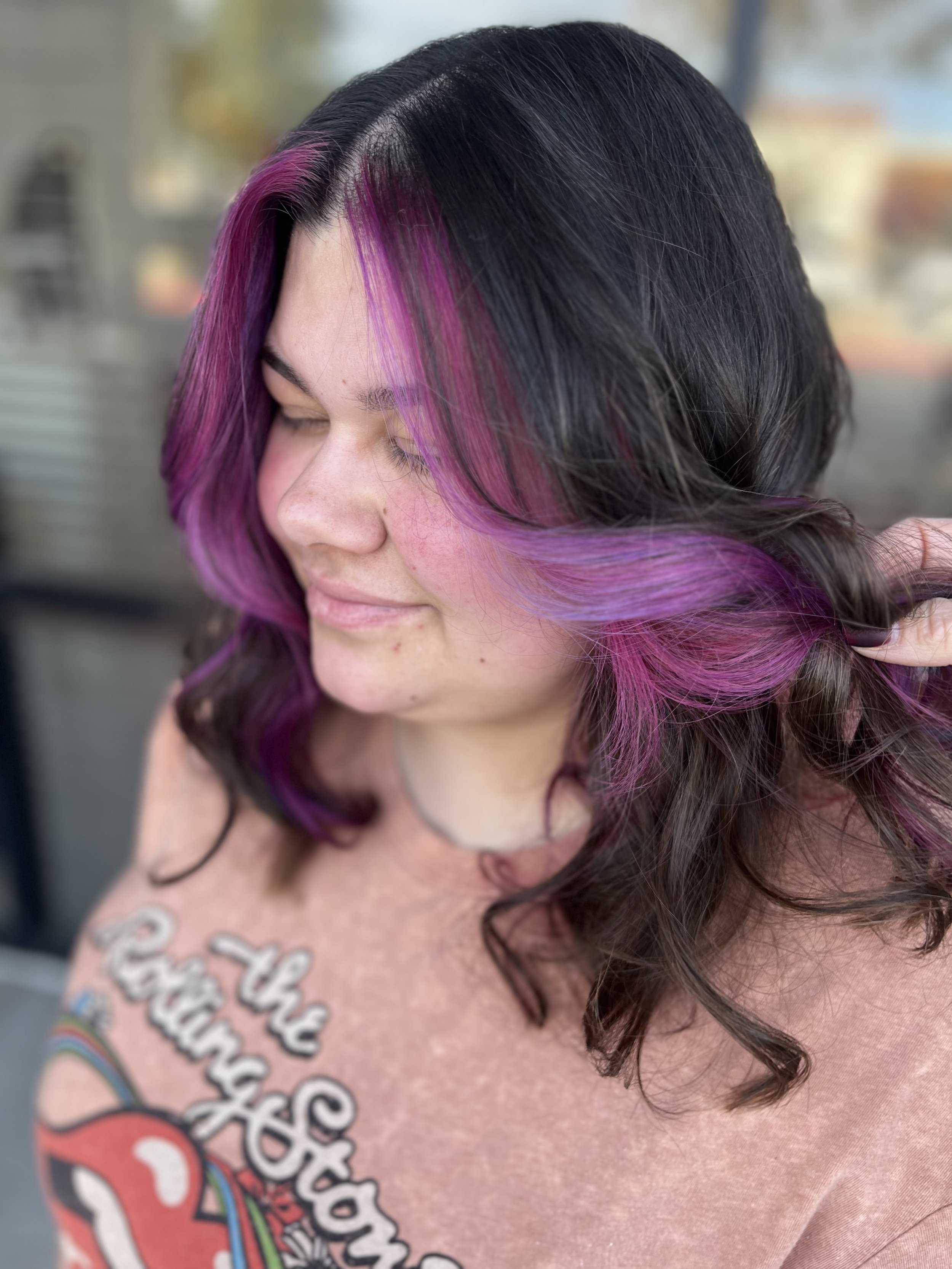 Woman with wavy dark hair highlighted with vibrant purple and pink streaks, smiling softly with her eyes closed, standing outdoors.