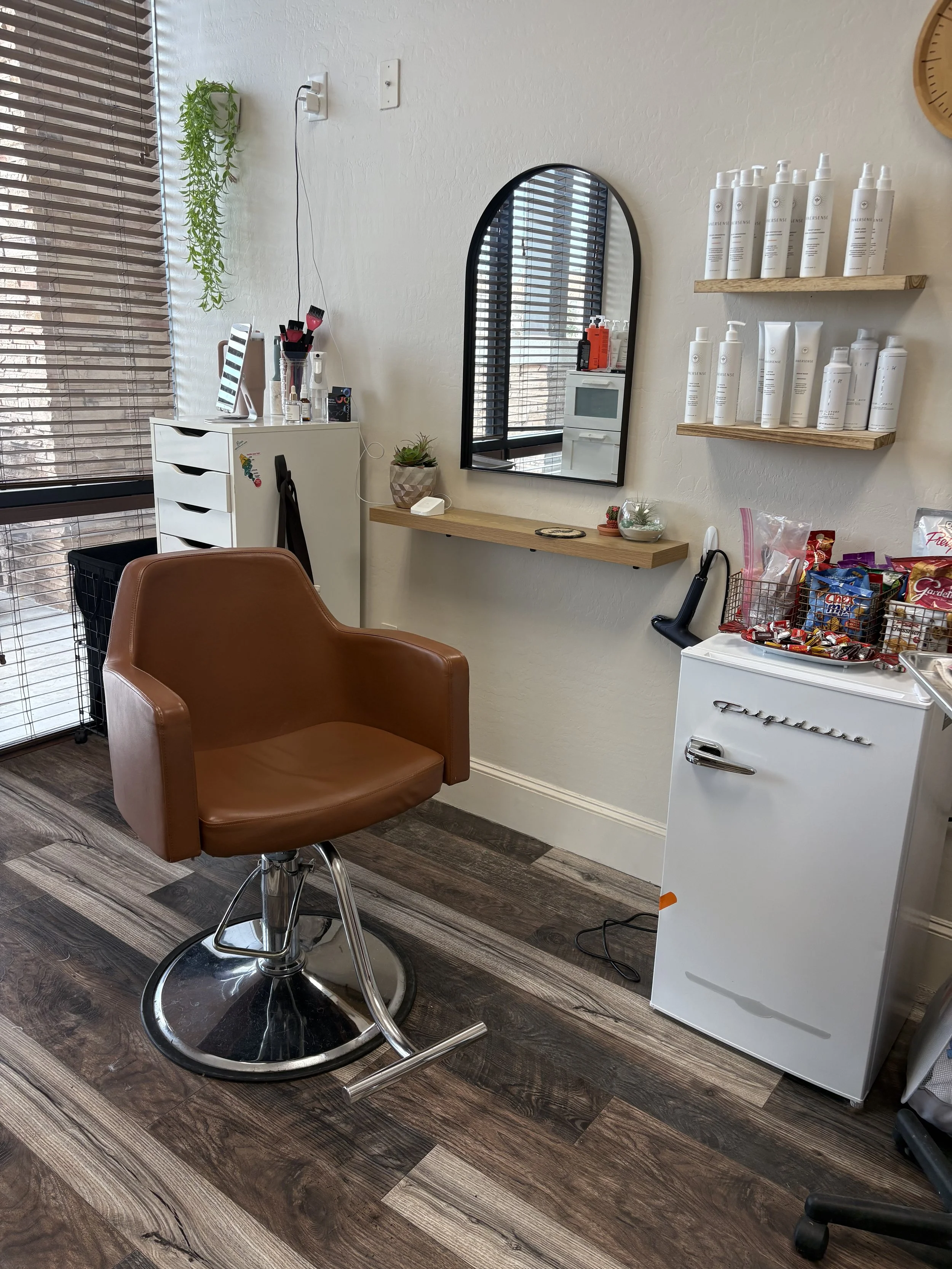 A hair salon station with a brown swivel chair, a mirror on the wall, and shelves with hair products. There is a small black basket and a white mini fridge with snacks on top, near a window with blinds. The room has wood flooring and a clock on the wall.