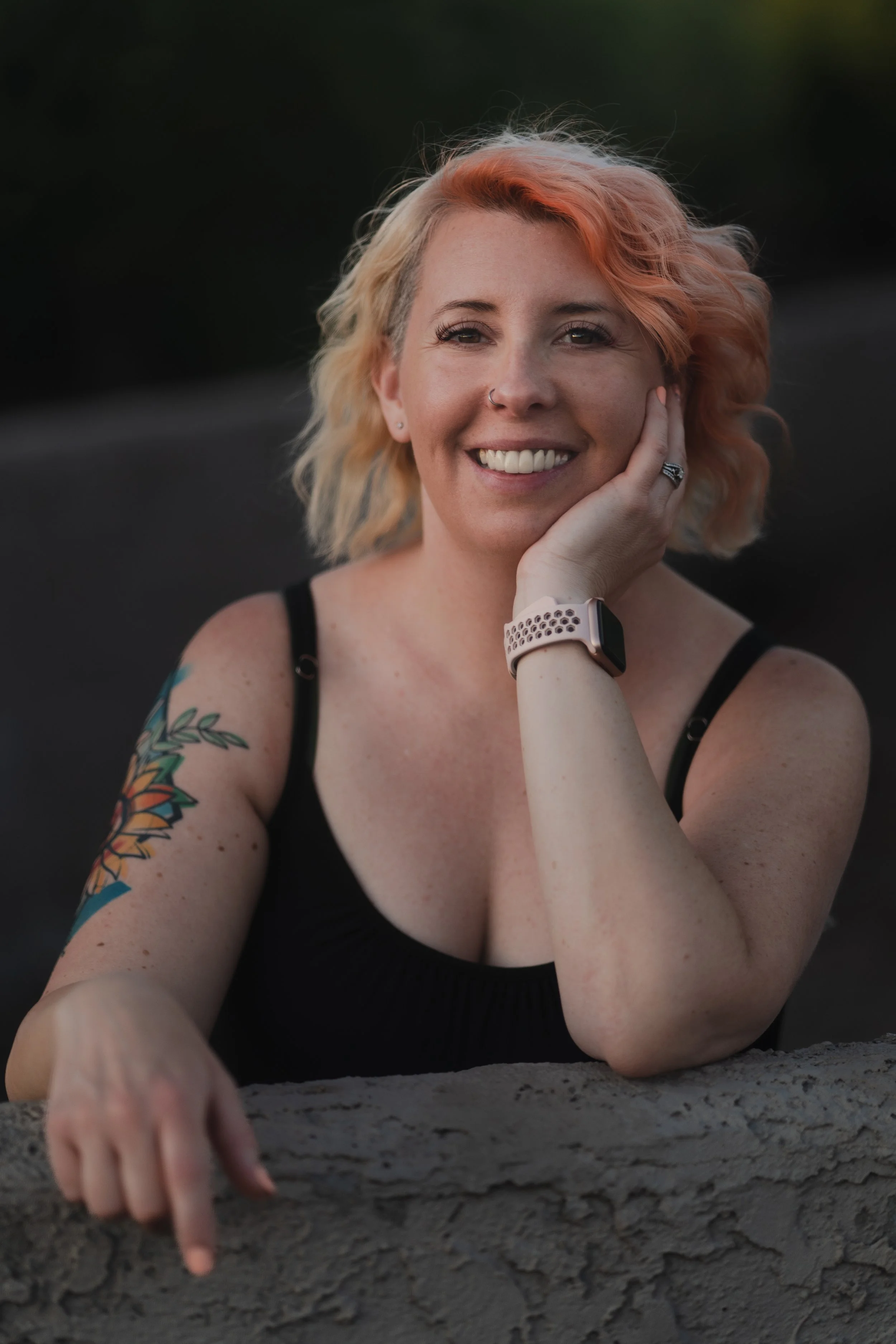 A smiling woman with blonde, pink, and orange hair, wearing a black top, seated outdoors, with a tattoo of a colorful butterfly on her arm, a smartwatch on her wrist, and resting her face on her hand.