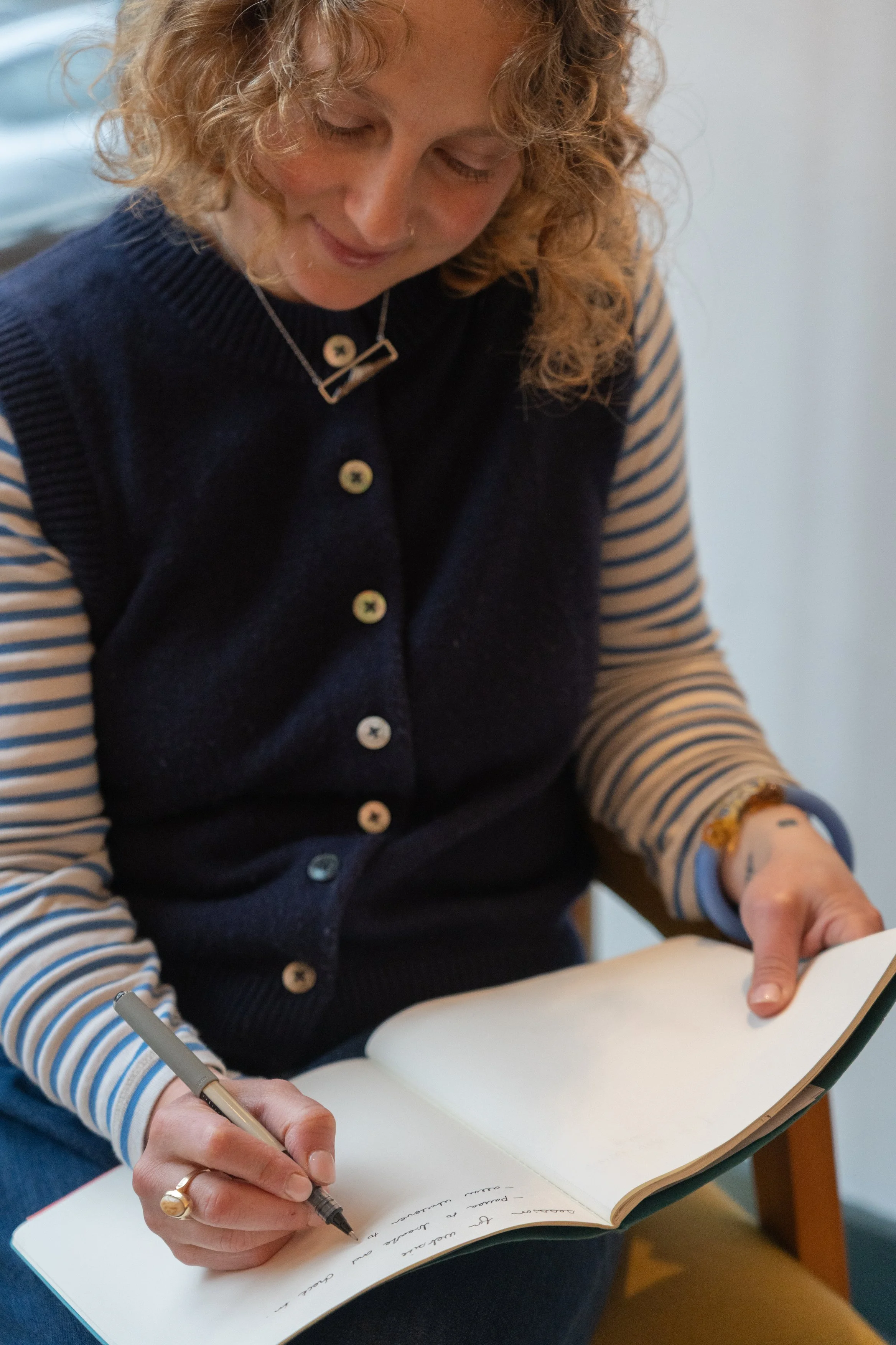 A woman with curly blonde hair is sitting and writing in a notebook with a pen. She is wearing a striped long-sleeve shirt underneath a navy blue vest, and has a bracelet and ring on her left hand.