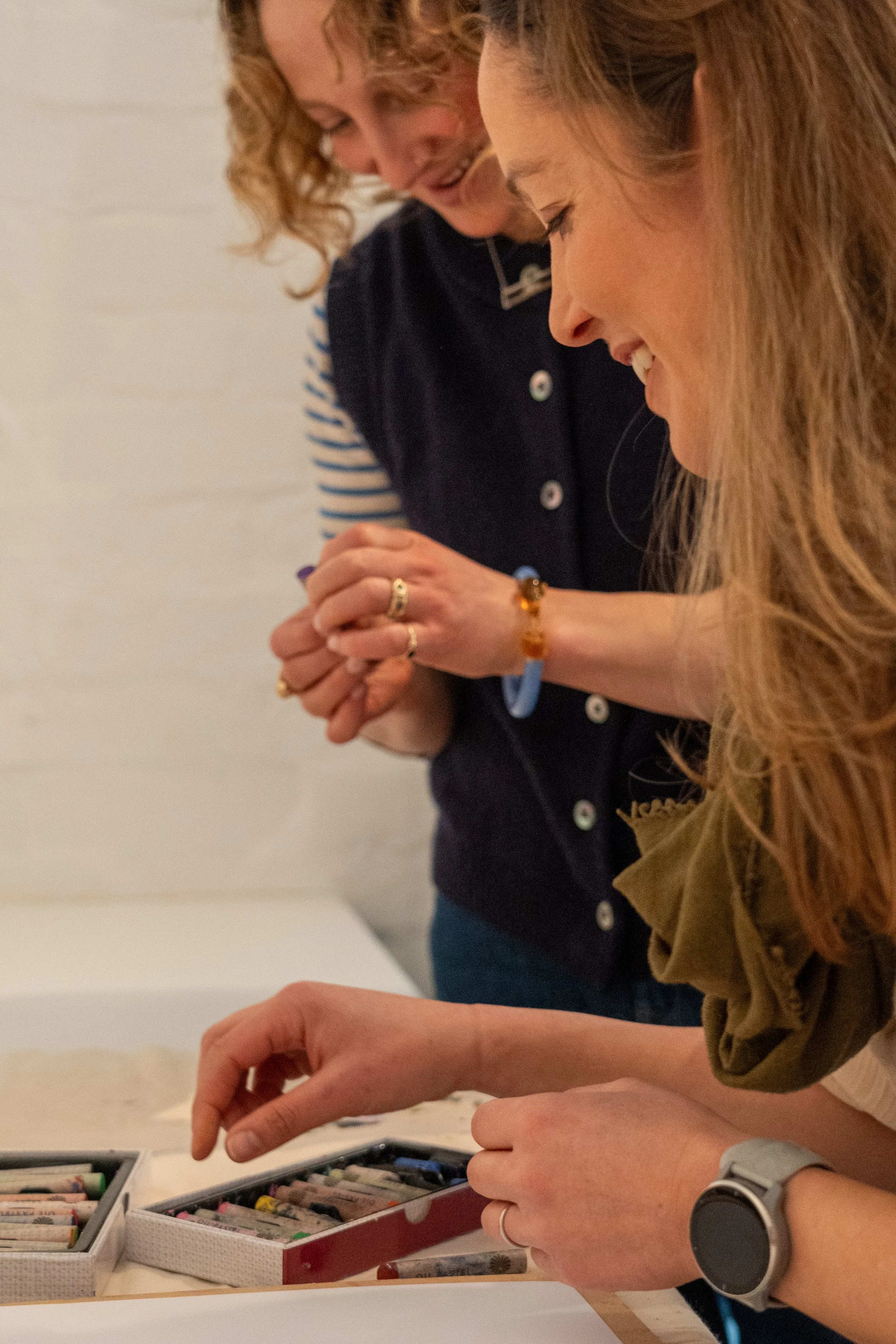 Two women smiling and looking at a collection of crayons or pastels in trays on a table.