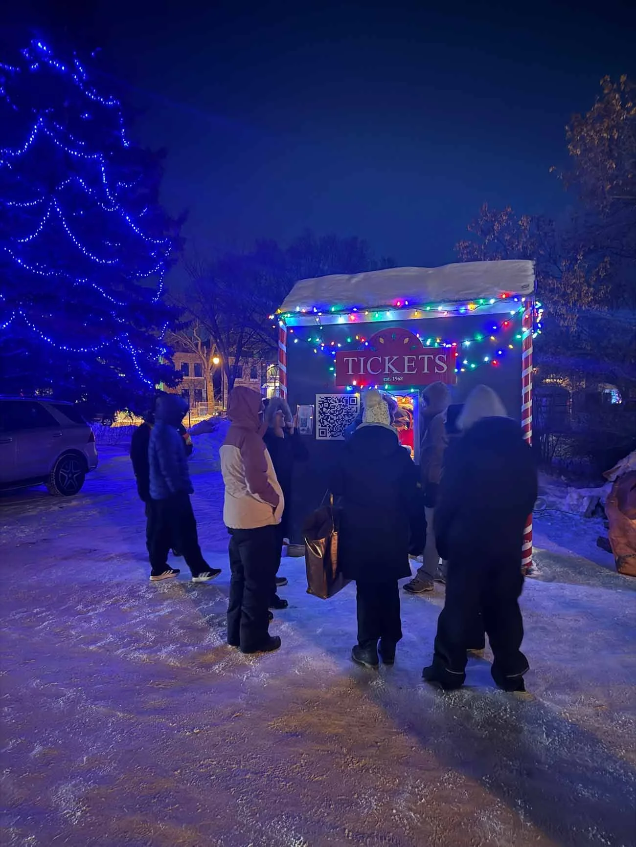 People standing in line at a ticket booth decorated with colorful Christmas lights, with a snowy ground and trees lit with blue lights in the background.