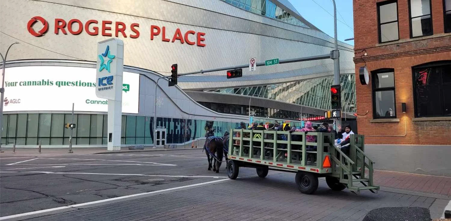 A horse hitsched to a wagon filled with bags and items, parked in front of Rogers Place arena, with street signs for 104th street and traffic lights visible.