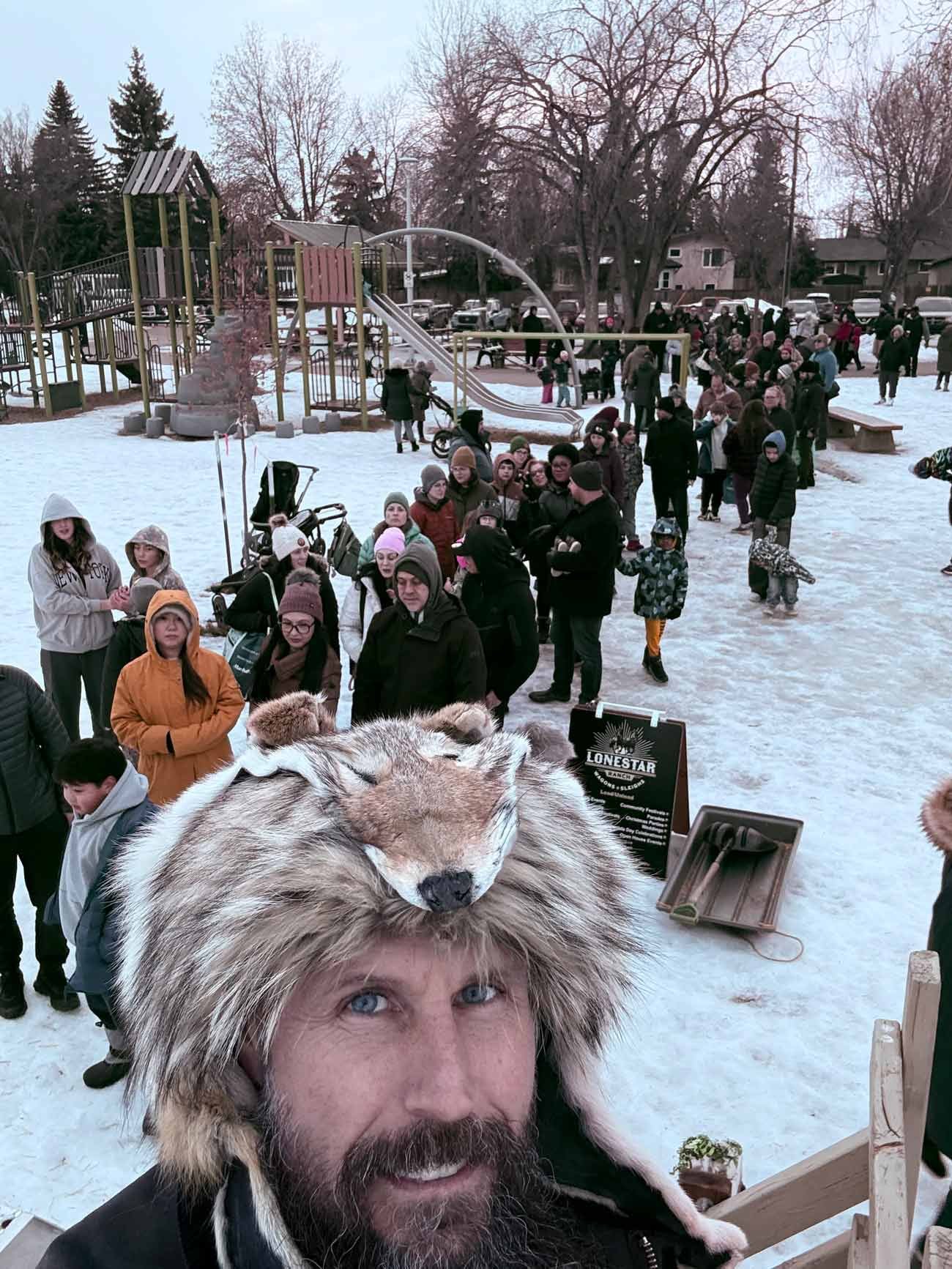 A man with a fur hat featuring a fox face, taking a selfie at a snowy outdoor event with children and adults in line, a playground in the background, and trees and buildings.